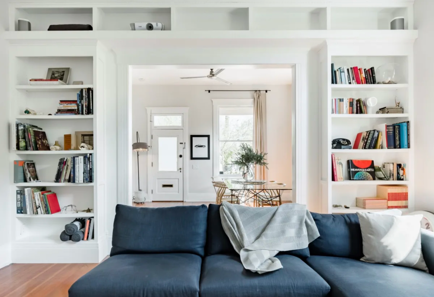 Living room with built-in white bookshelves, a navy sofa with a light-colored throw blanket and pillow, visible hardwood floor, and a view into a dining area with a table, chairs, and windows in the background.