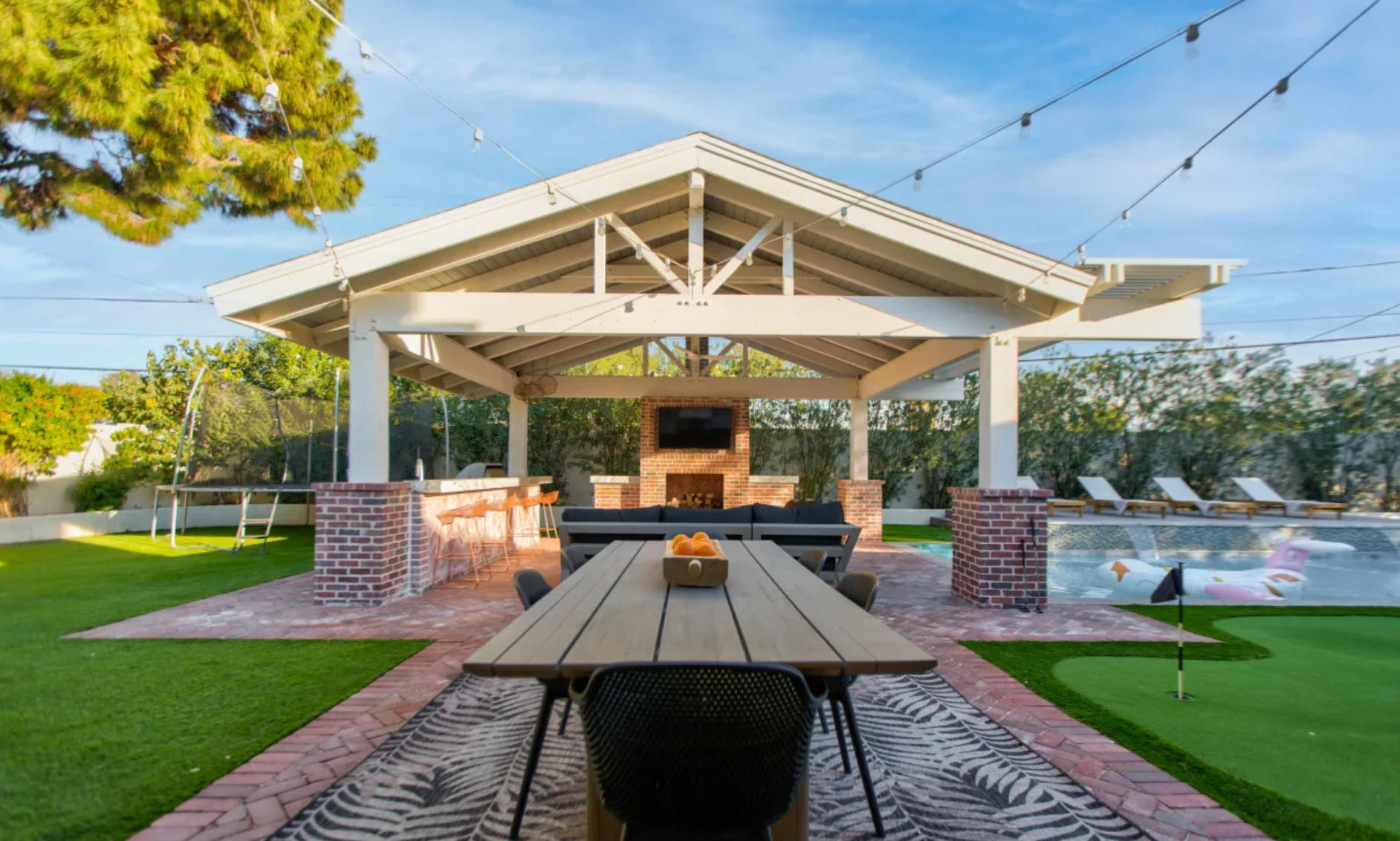 A backyard patio area with a wooden dining table, black chairs, and an outdoor fireplace with a television mounted above it under a white pergola. There is a pool with floating toys, a trampoline, and lounge chairs with umbrellas, surrounded by green grass and trees, under a blue sky.