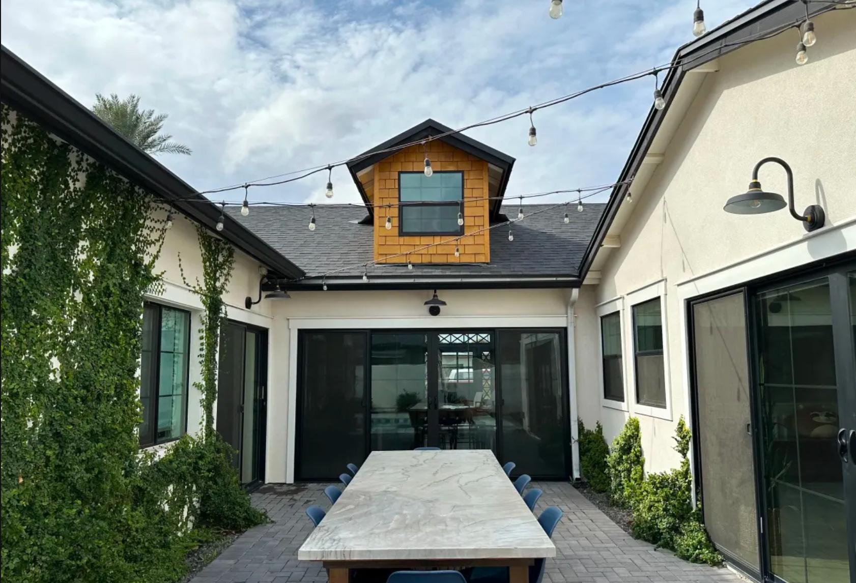 Outdoor patio with a long dining table, surrounded by blue chairs, enclosed by a white house with large glass sliding doors and windows. String lights hang overhead, with a blue sky overhead and some greenery on the left and right sides.
