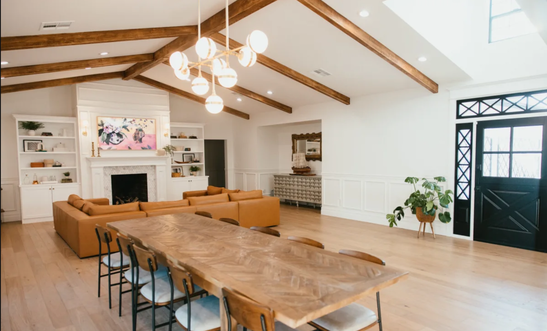 Spacious living room with high vaulted ceiling featuring wooden beams, a modern chandelier, a fireplace with built-in shelves, tan sectional sofa, wooden dining table with chairs, potted plant near black door, and white walls with wainscoting.