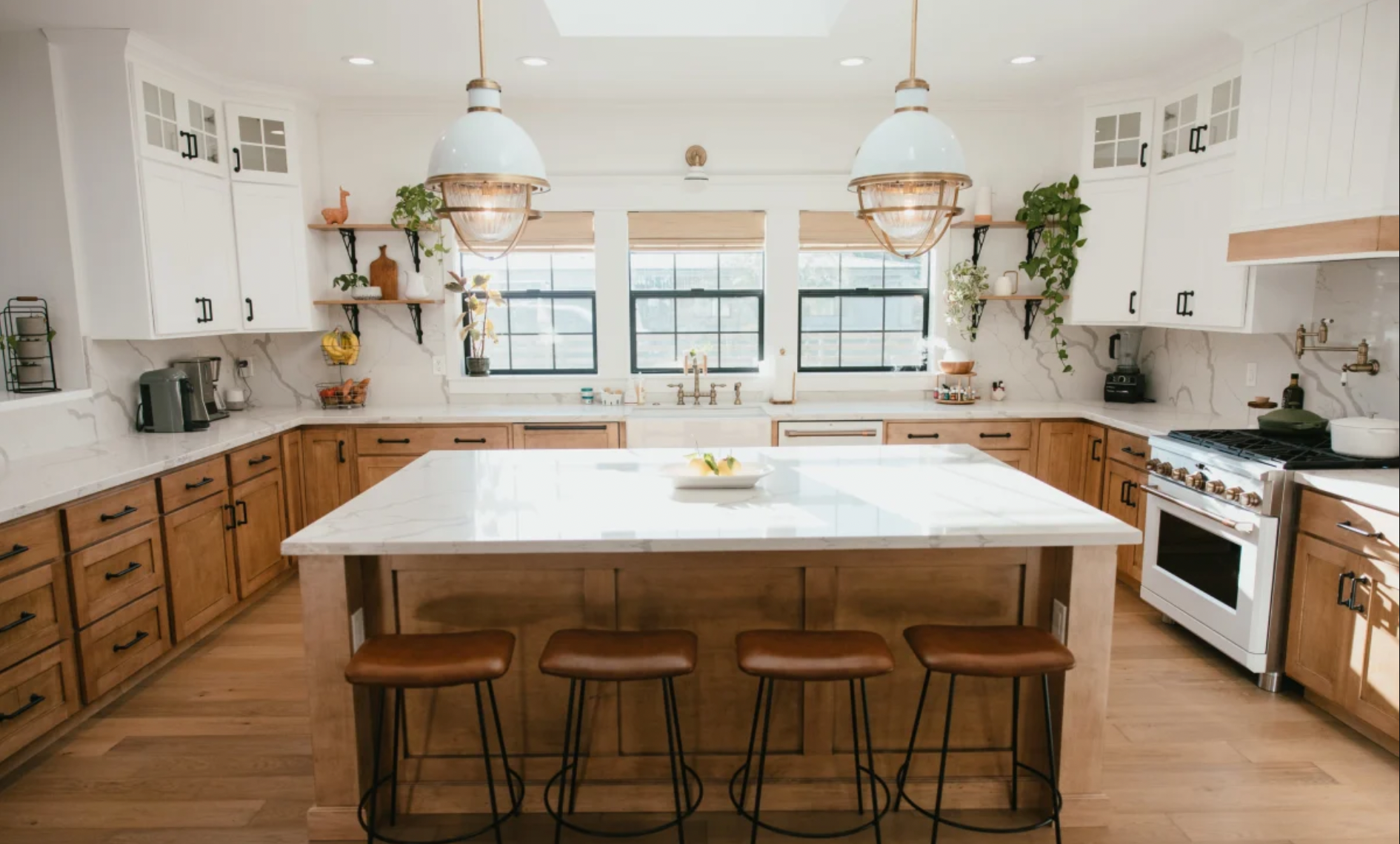 Modern kitchen with white upper cabinets, wood lower cabinets, a large marble island, and three brown stools. Features include pendant lighting, black hardware, windows behind the sink, and decor with plants and pottery.