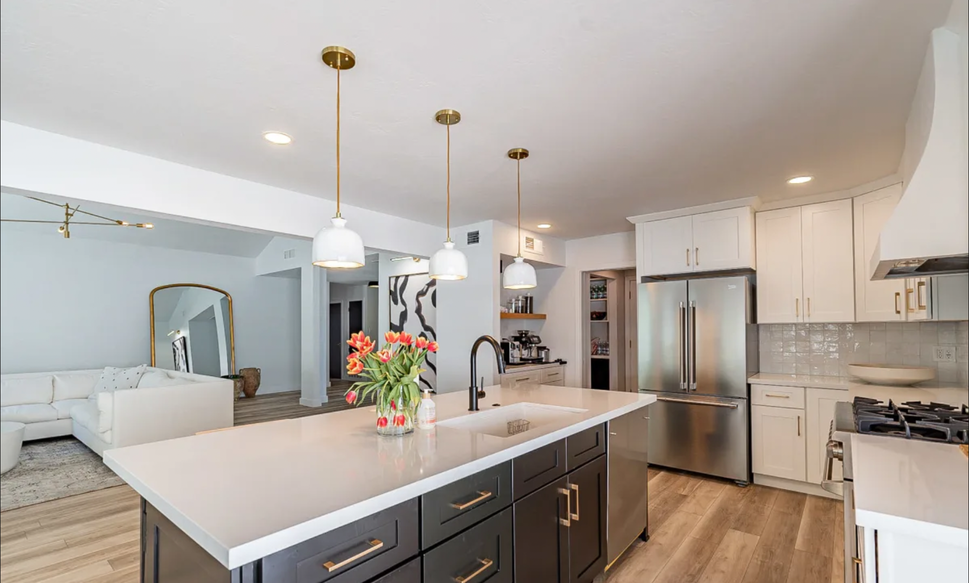 Modern kitchen with white cabinetry, stainless steel appliances, black island with gold hardware, three white pendant lights, and a vase of red tulips on the island. Adjacent living room with white sofa and large mirror.