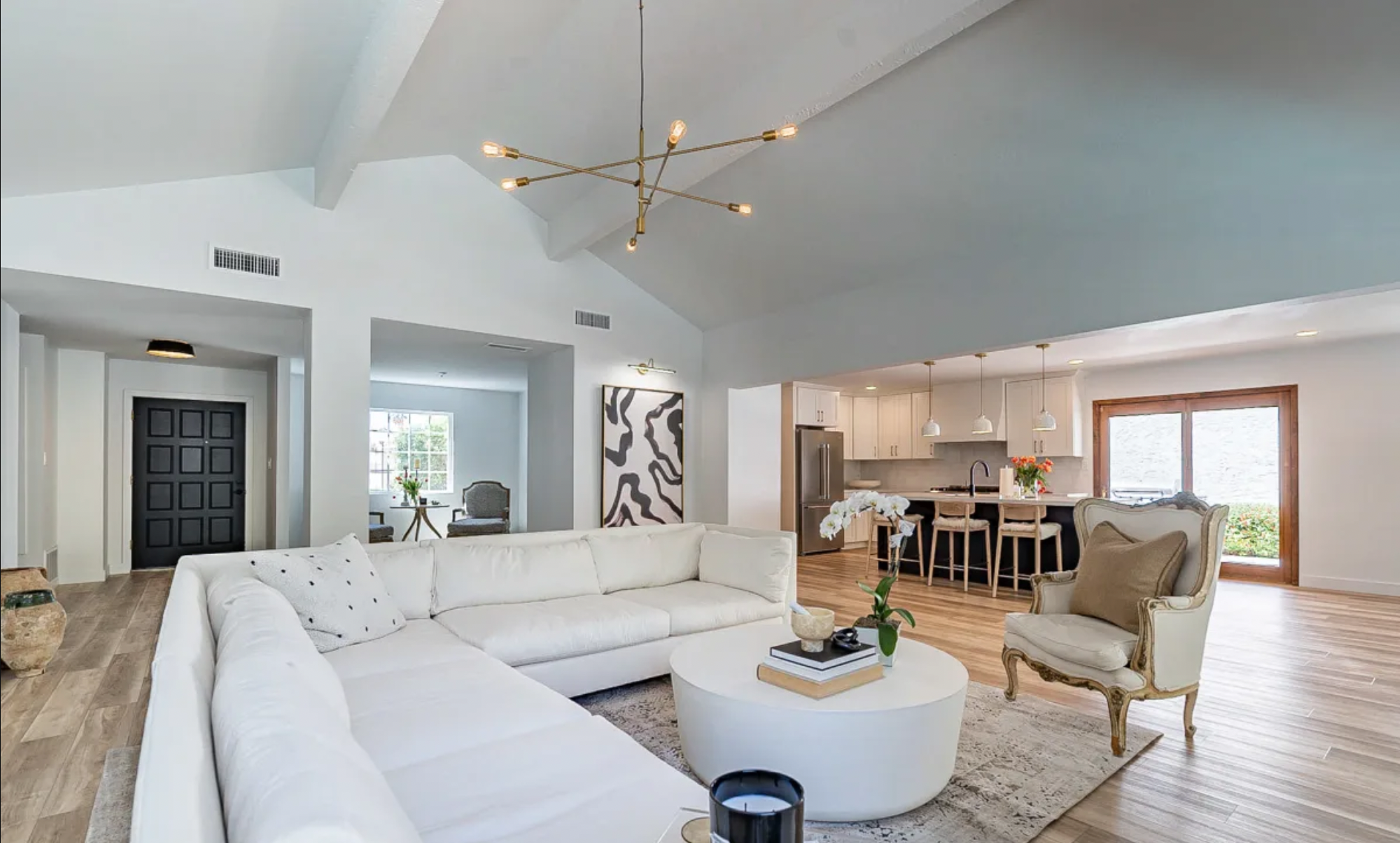 Open-concept living room with white sectional sofa, round coffee table, and vintage armchair, adjacent to a kitchen with white cabinets and barstools, decorated with modern art and flowers.