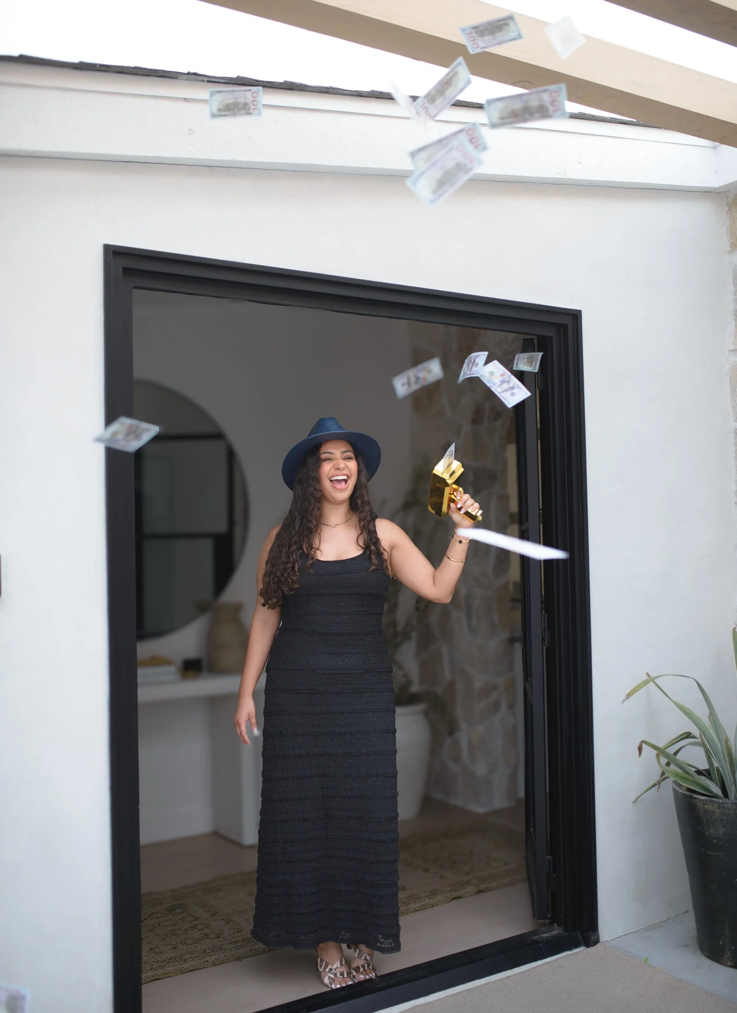 A woman standing in a doorway, celebrating and smiling, with money falling around her, holding a small golden hammer.