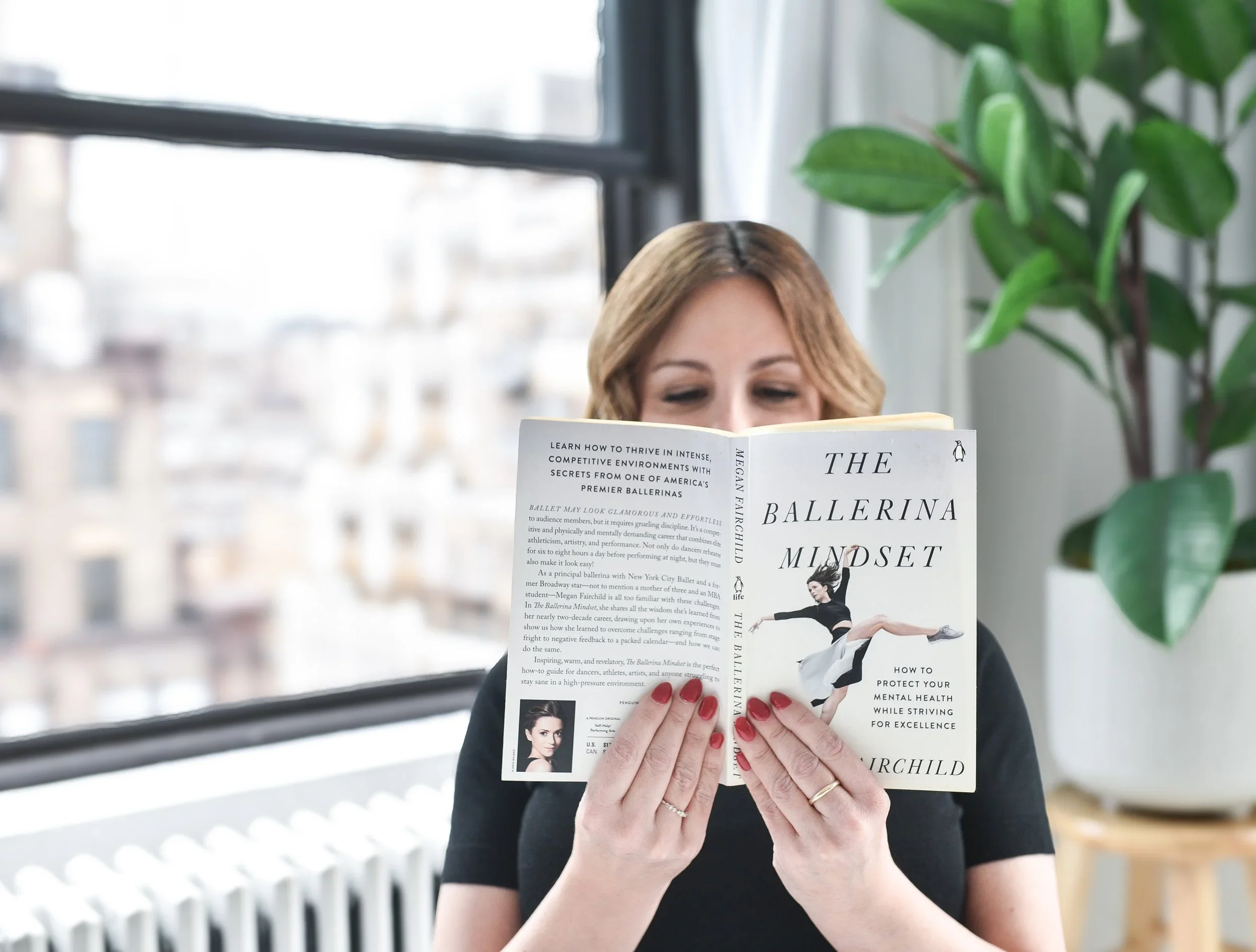 A woman with light brown hair and red nail polish, wearing a black shirt, is reading a book titled 'The Ballerina Mindset' by Megan Fairchild. She is sitting near a window with a view of buildings outside, and there is a large green plant on her right.
