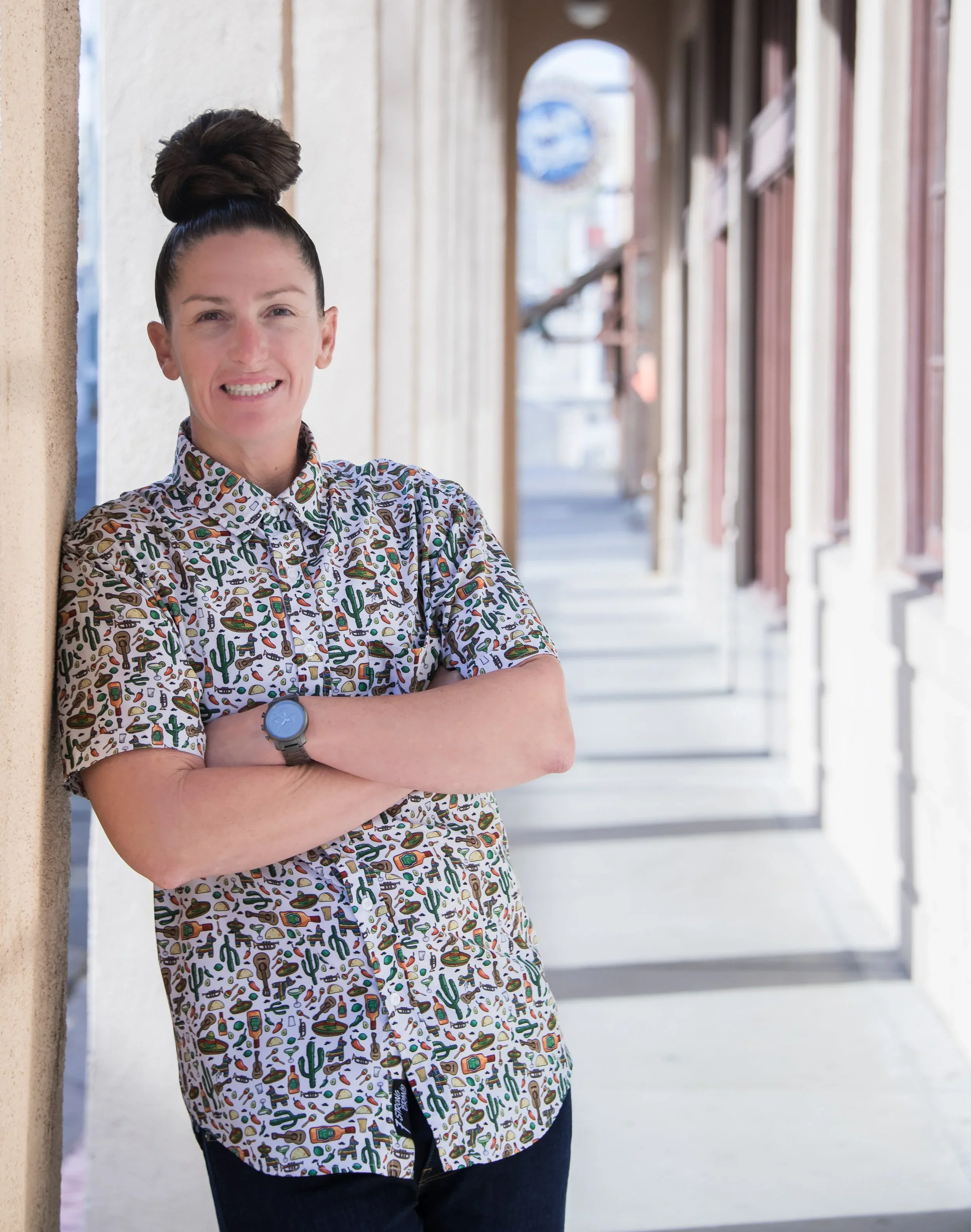 A woman stands with arms crossed, leaning against a wall outside a row of shops, smiling at the camera.