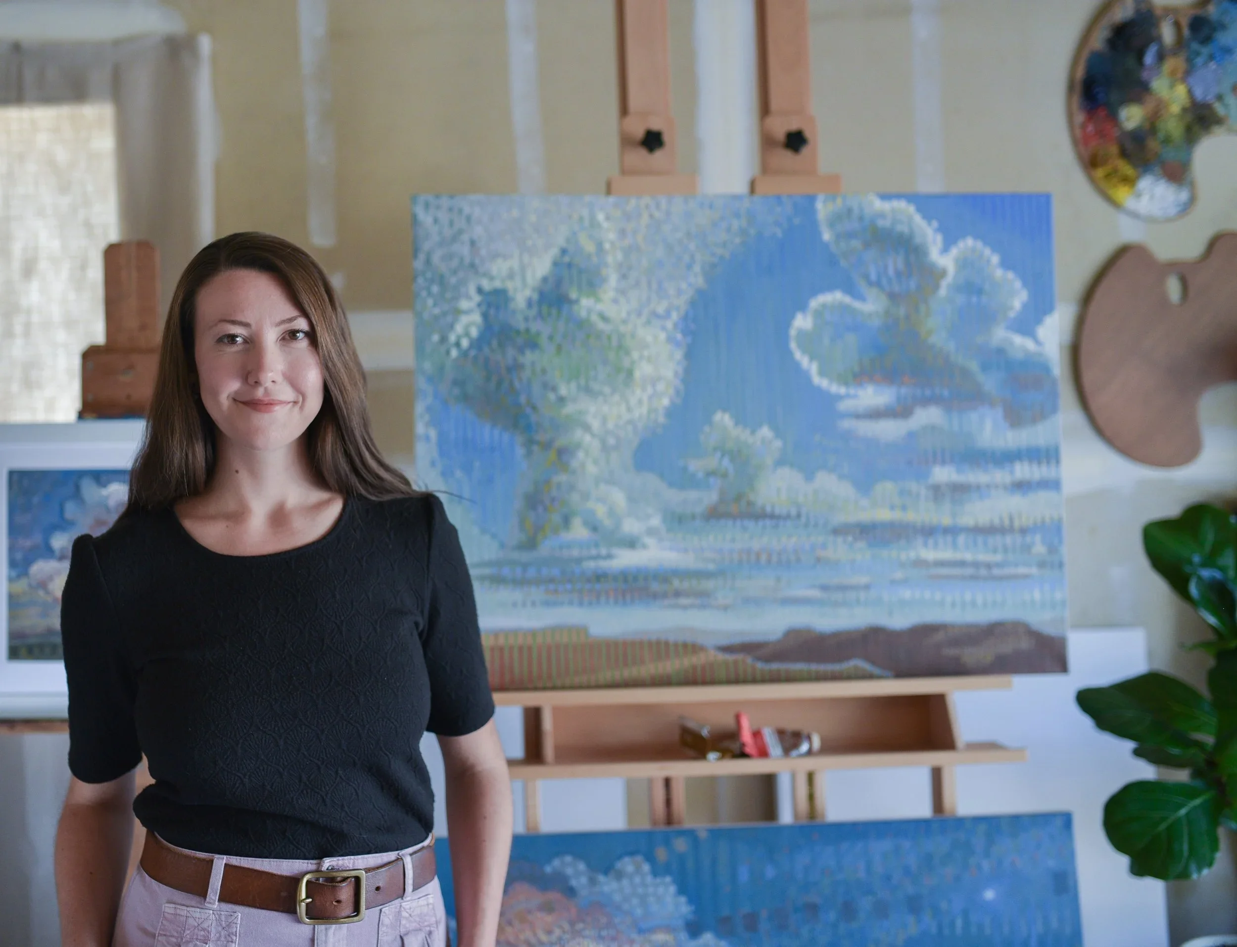 Young woman standing in art studio with paintings of clouds and sky in the background.