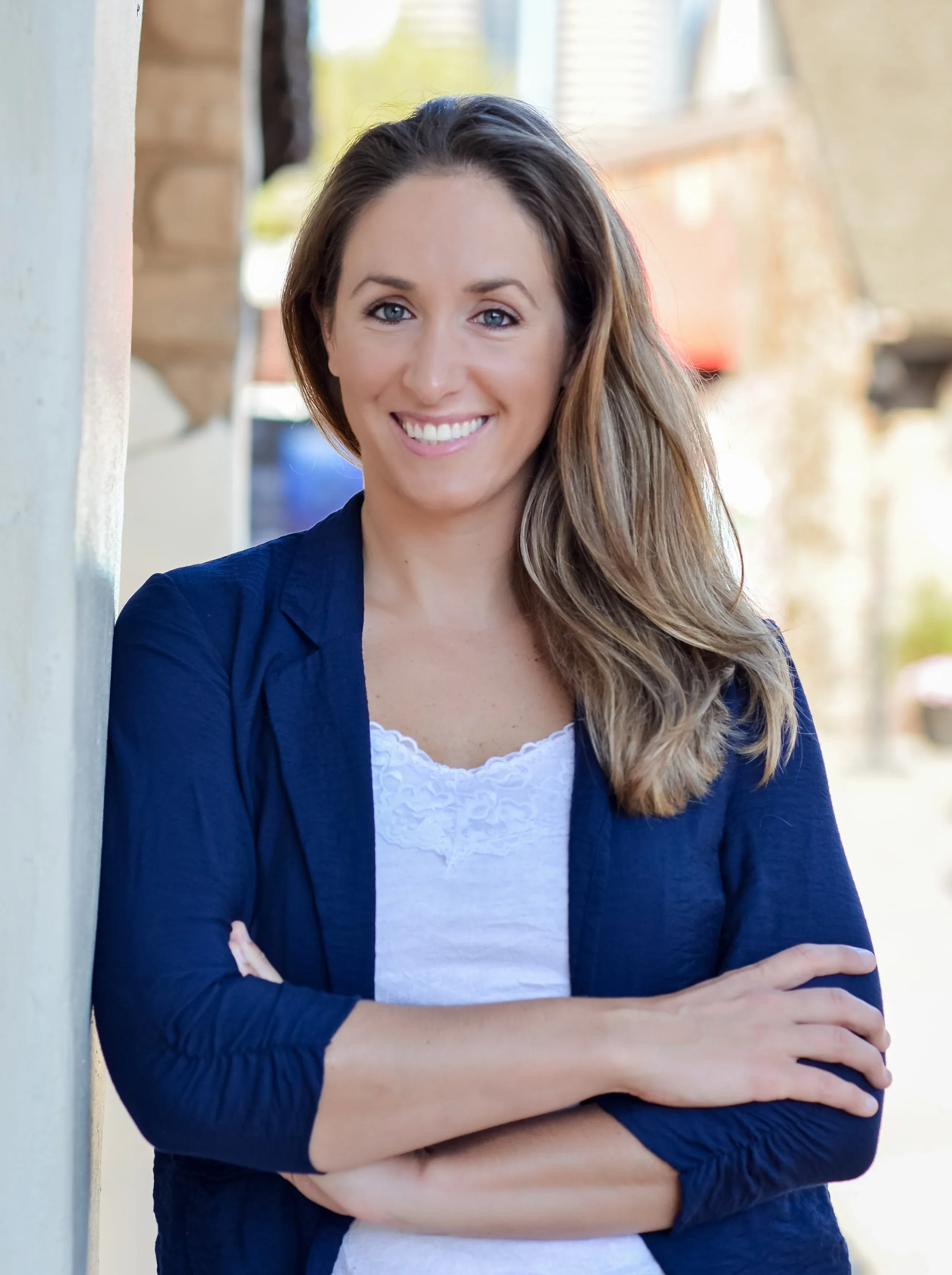 Young woman with long brown hair, wearing a navy blue blazer over a white top, smiling with arms crossed, standing outdoors.