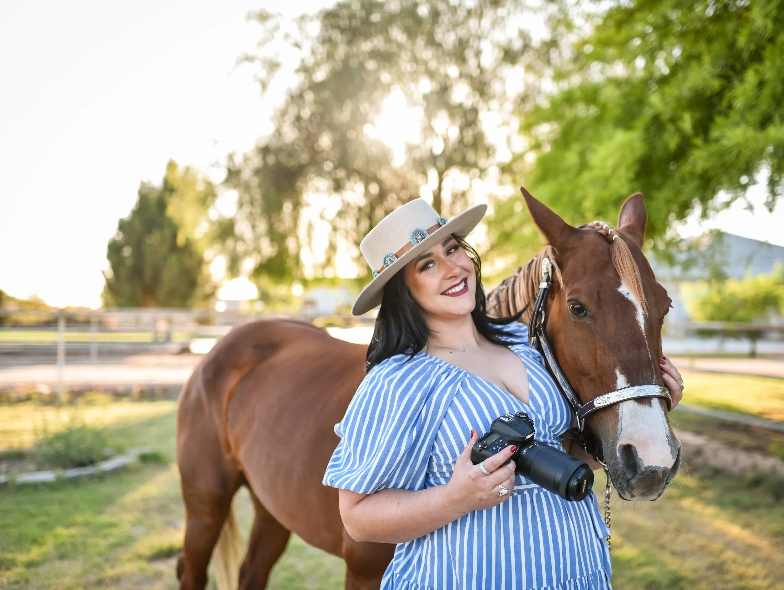 A woman in a blue and white striped dress holding a camera and smiling while standing beside a brown horse outside in a green, sunlit field.