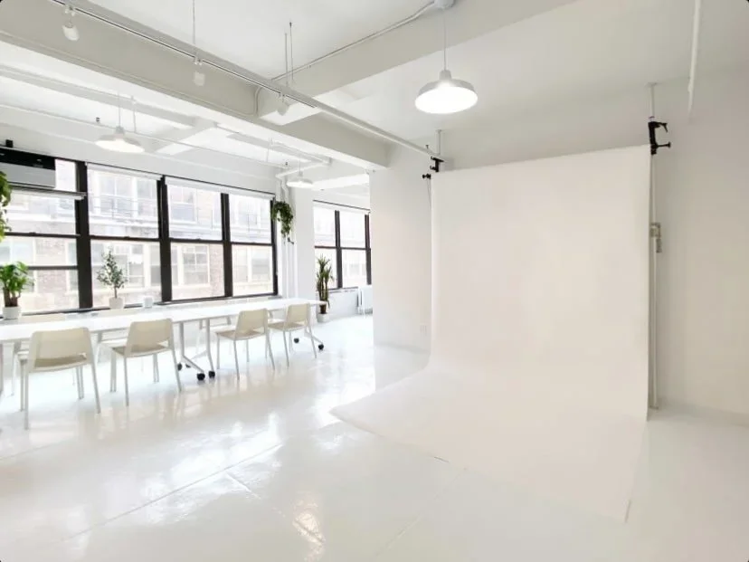 Bright industrial-style photo studio with white backdrop, large windows, chairs, and potted plants.