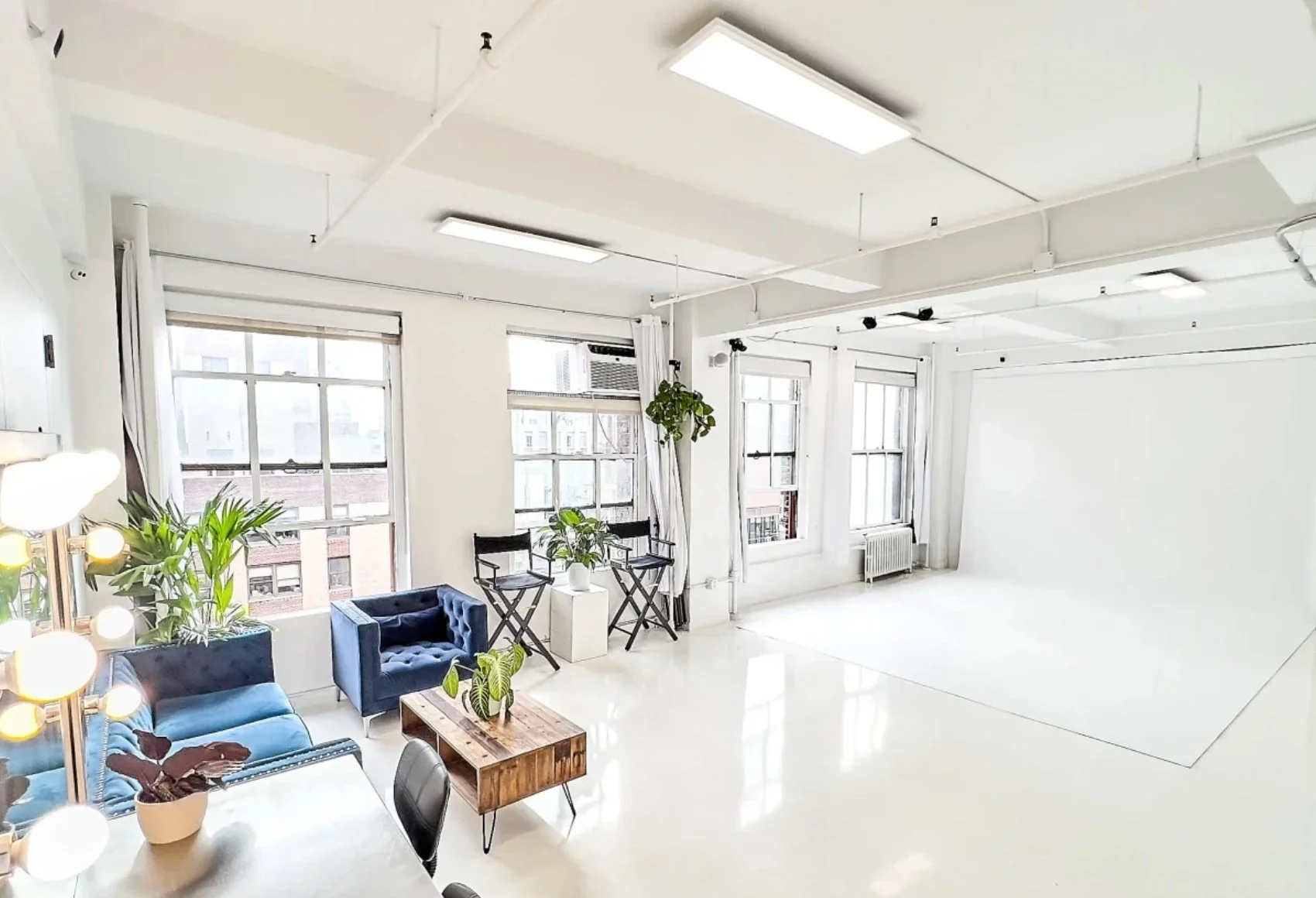 Bright studio with white walls, large windows, and minimalistic furniture including blue chairs, a wooden table, and potted plants.