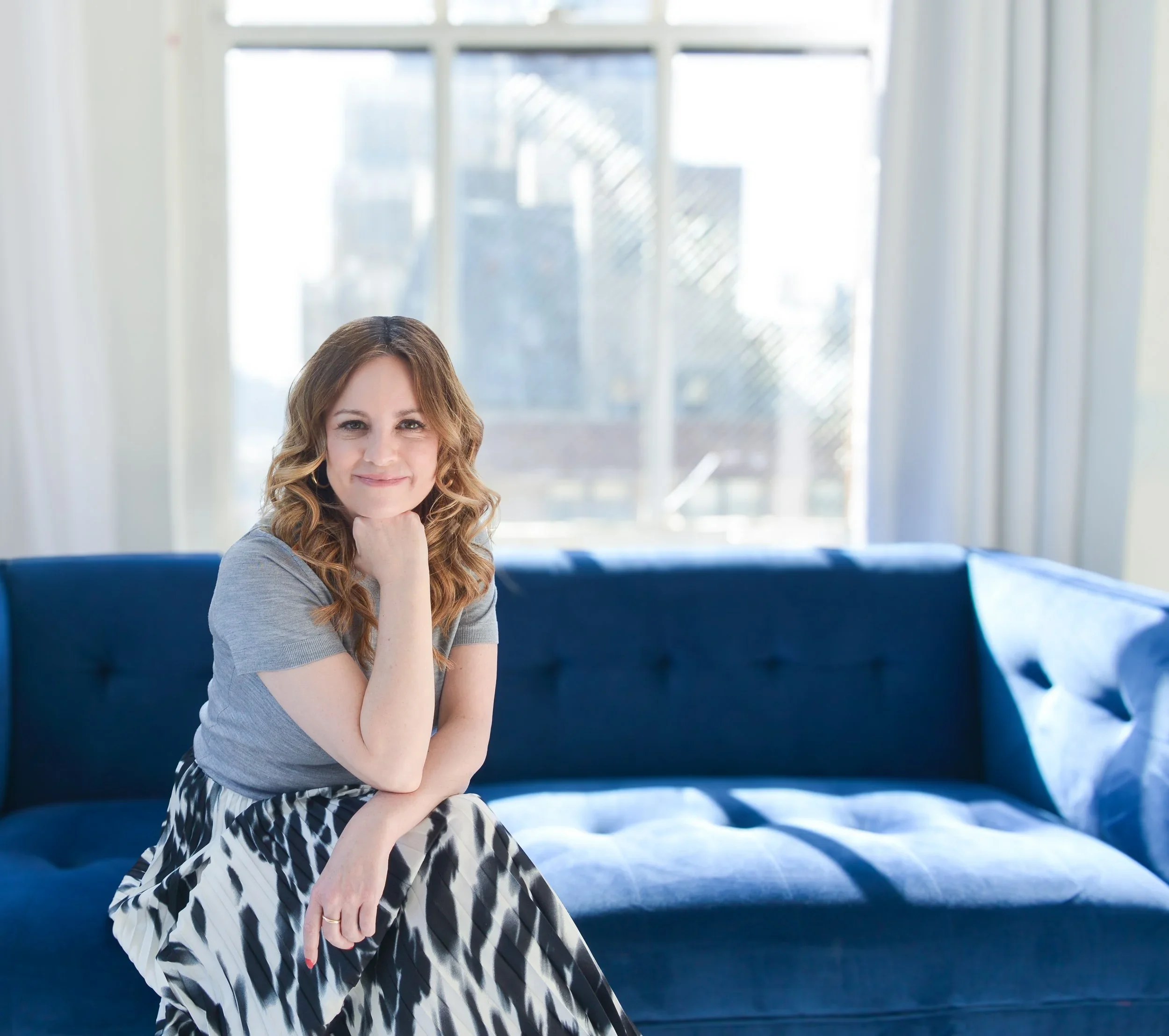 A woman with wavy brown hair sitting on a blue sofa in a well-lit room with large windows