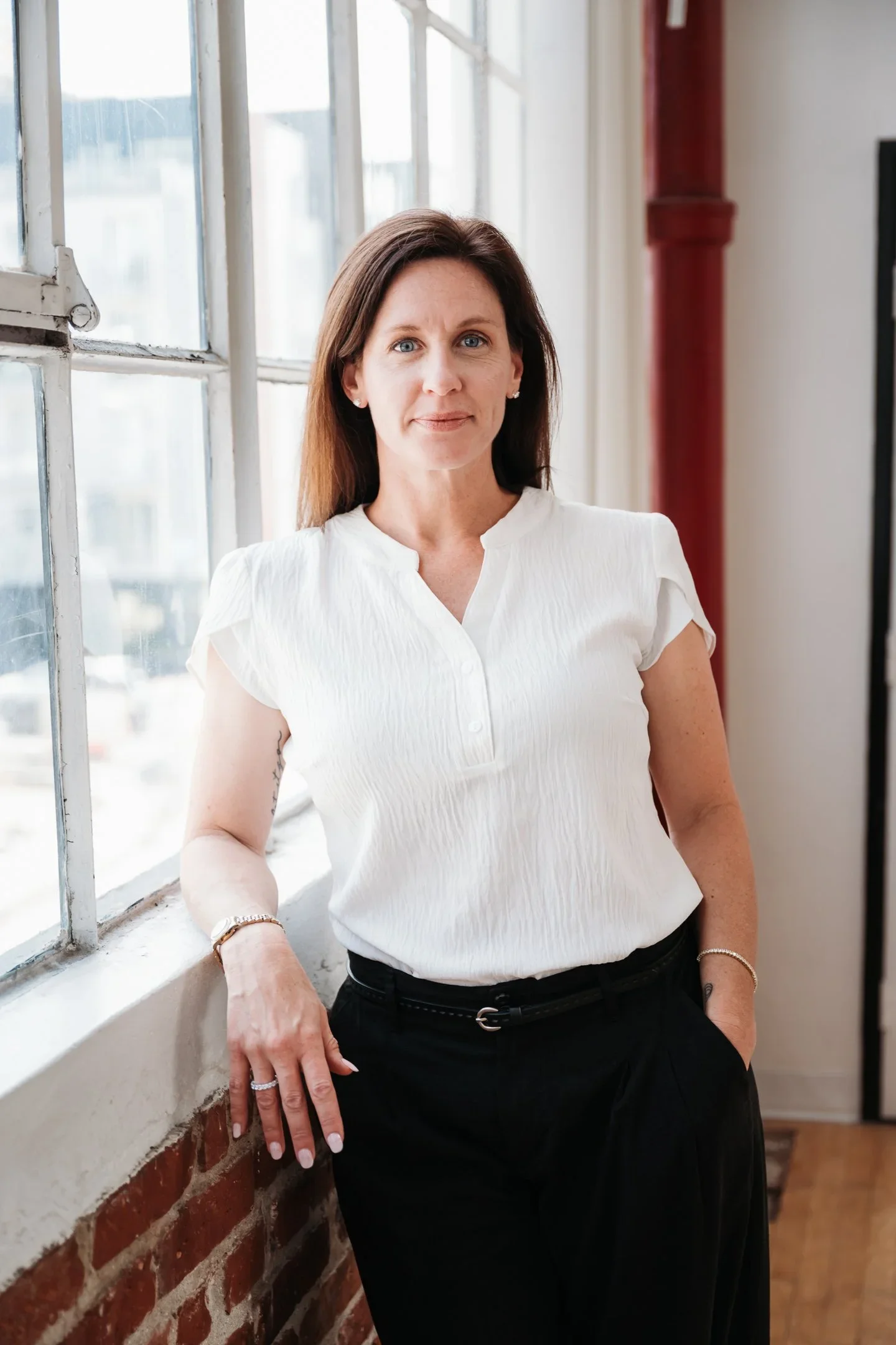 A woman with brown hair wearing a white blouse and black pants standing by a large window with a city view.