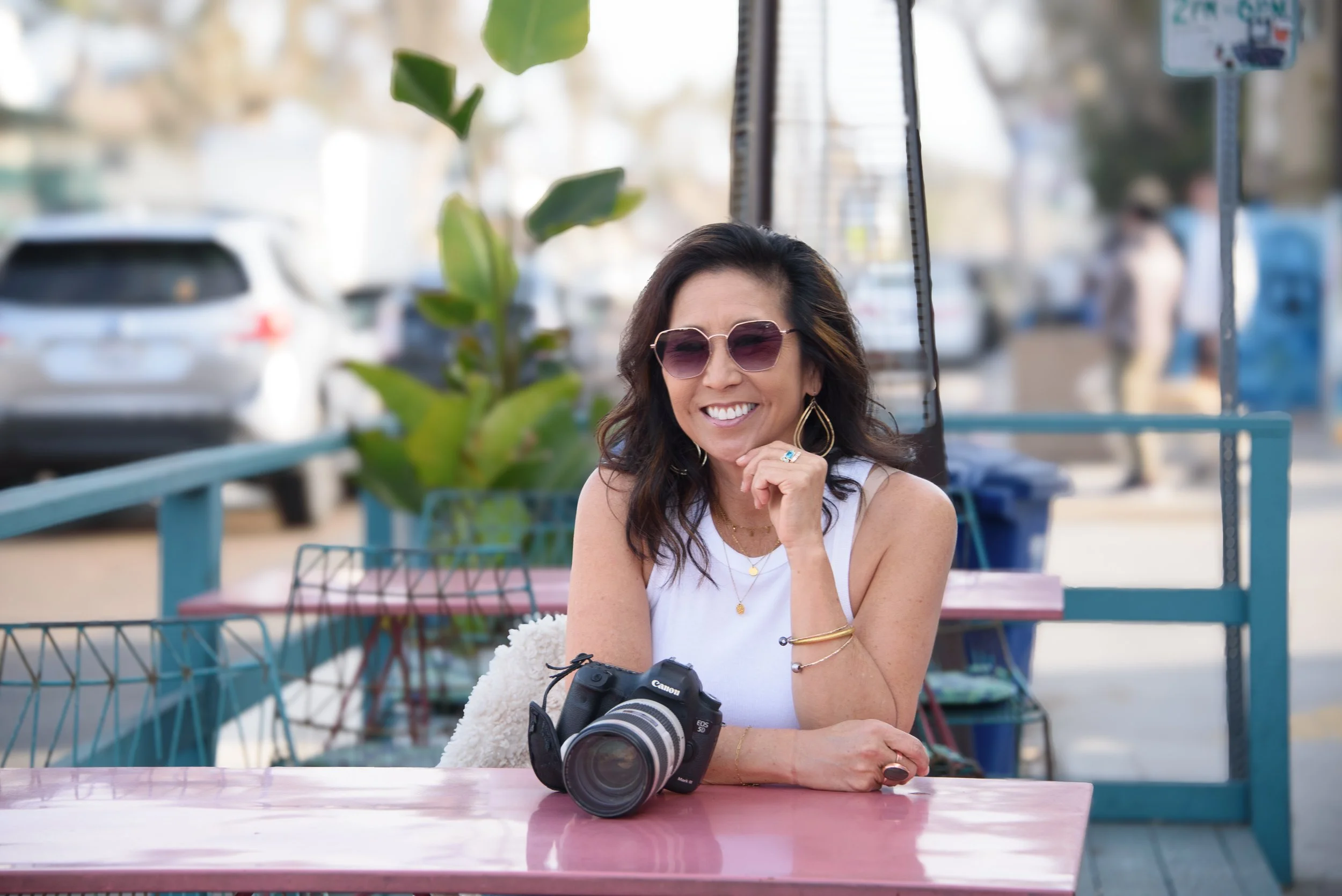 A smiling woman with dark hair, wearing sunglasses and jewelry, sitting at a pink table with a camera on it, outdoors during daytime.