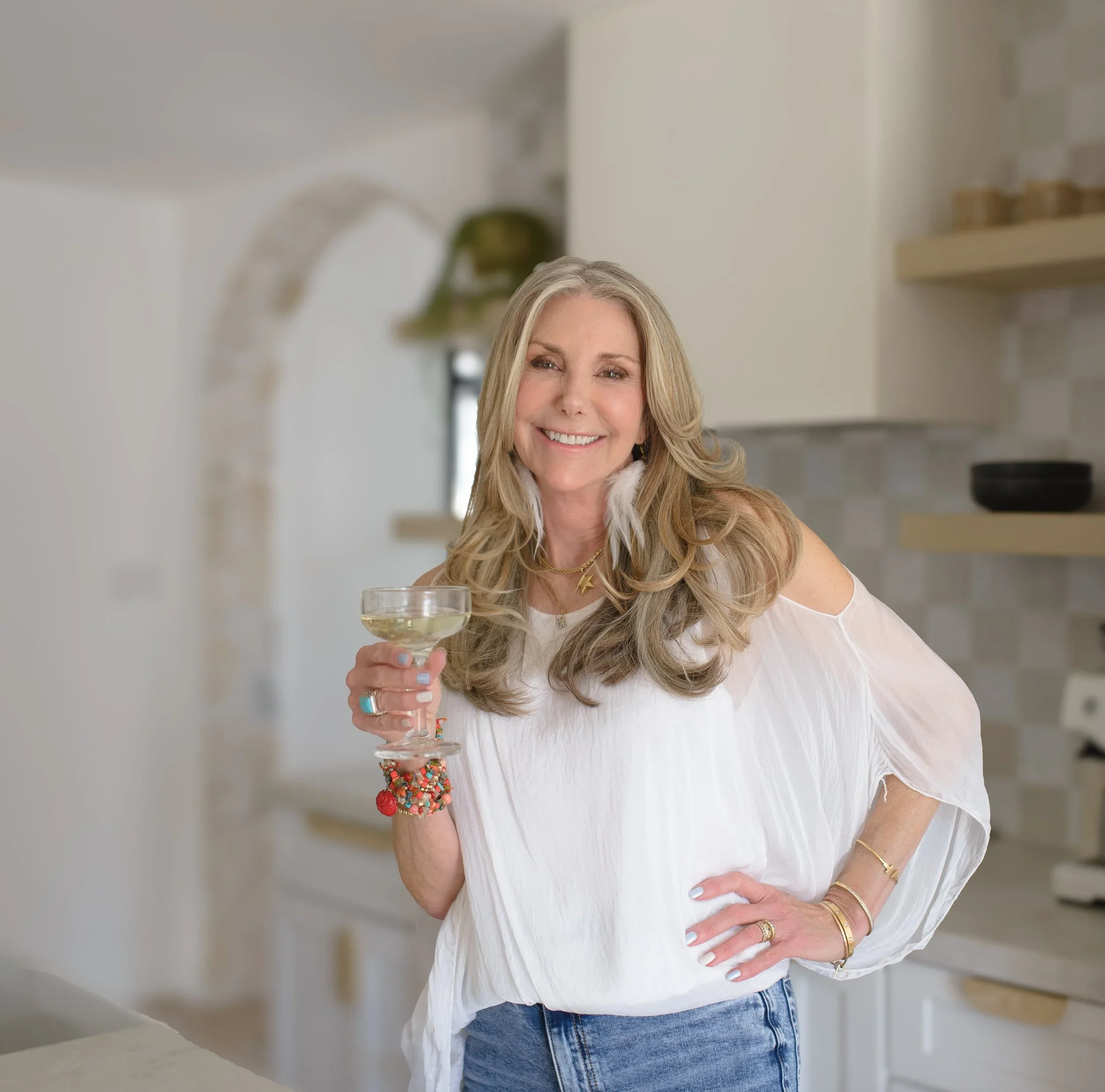 A woman smiling and holding a glass of white wine in a home kitchen.