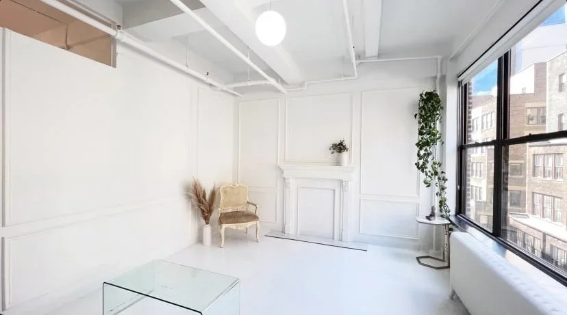 Bright, minimalist room with large windows, white walls, a vintage armchair, potted plants, and a glass coffee table.