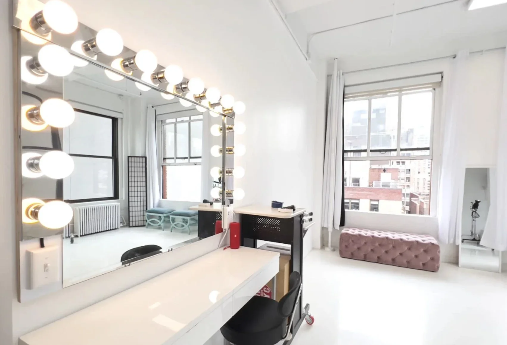 Bright makeup room with large mirror surrounded by globe vanity lights, black styling chair, and window with city view.