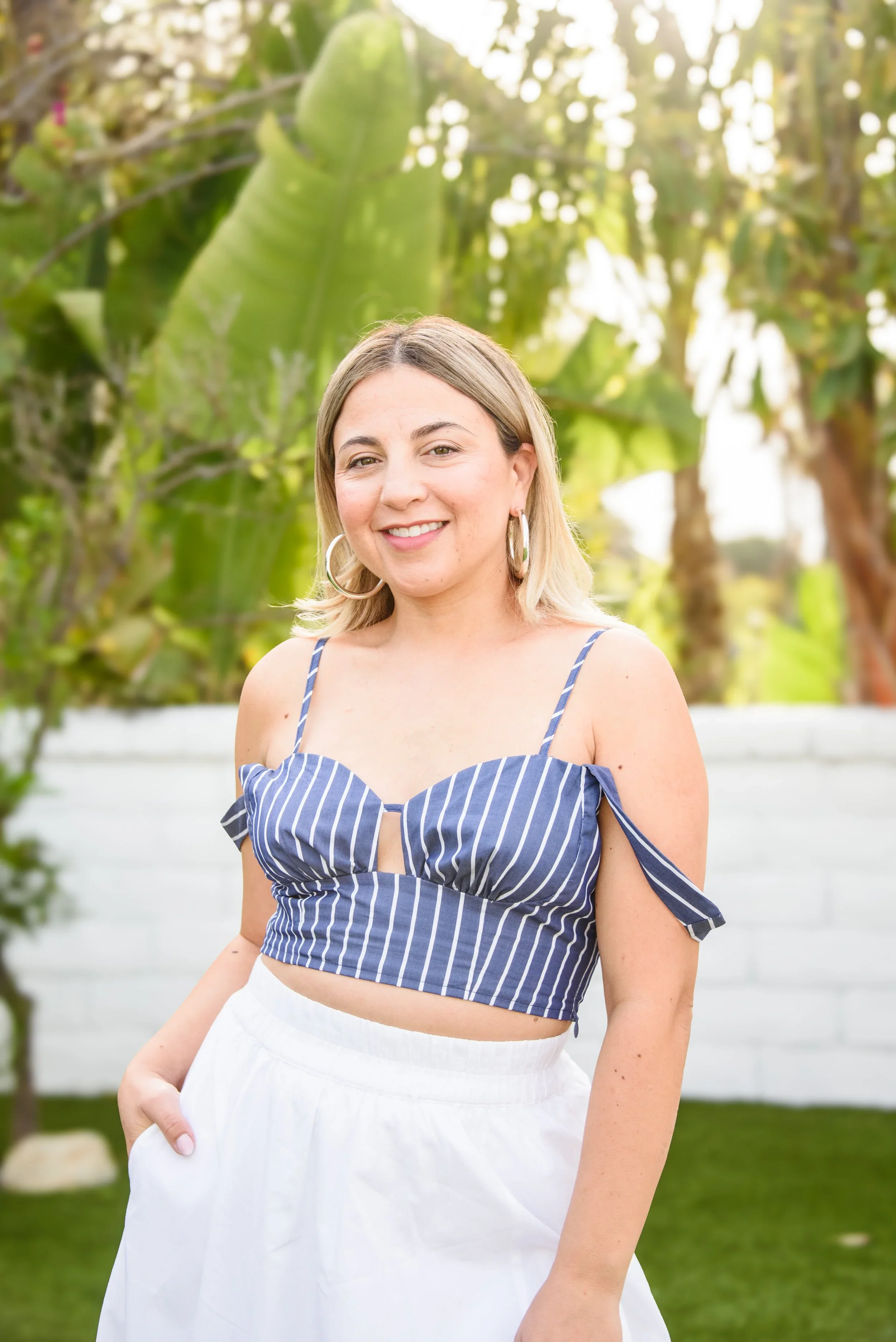 A woman with blonde hair and hoop earrings, wearing a blue and white striped cropped top with cold shoulder sleeves and white high-waisted pants, standing outdoors in front of green plants and trees, smiling.