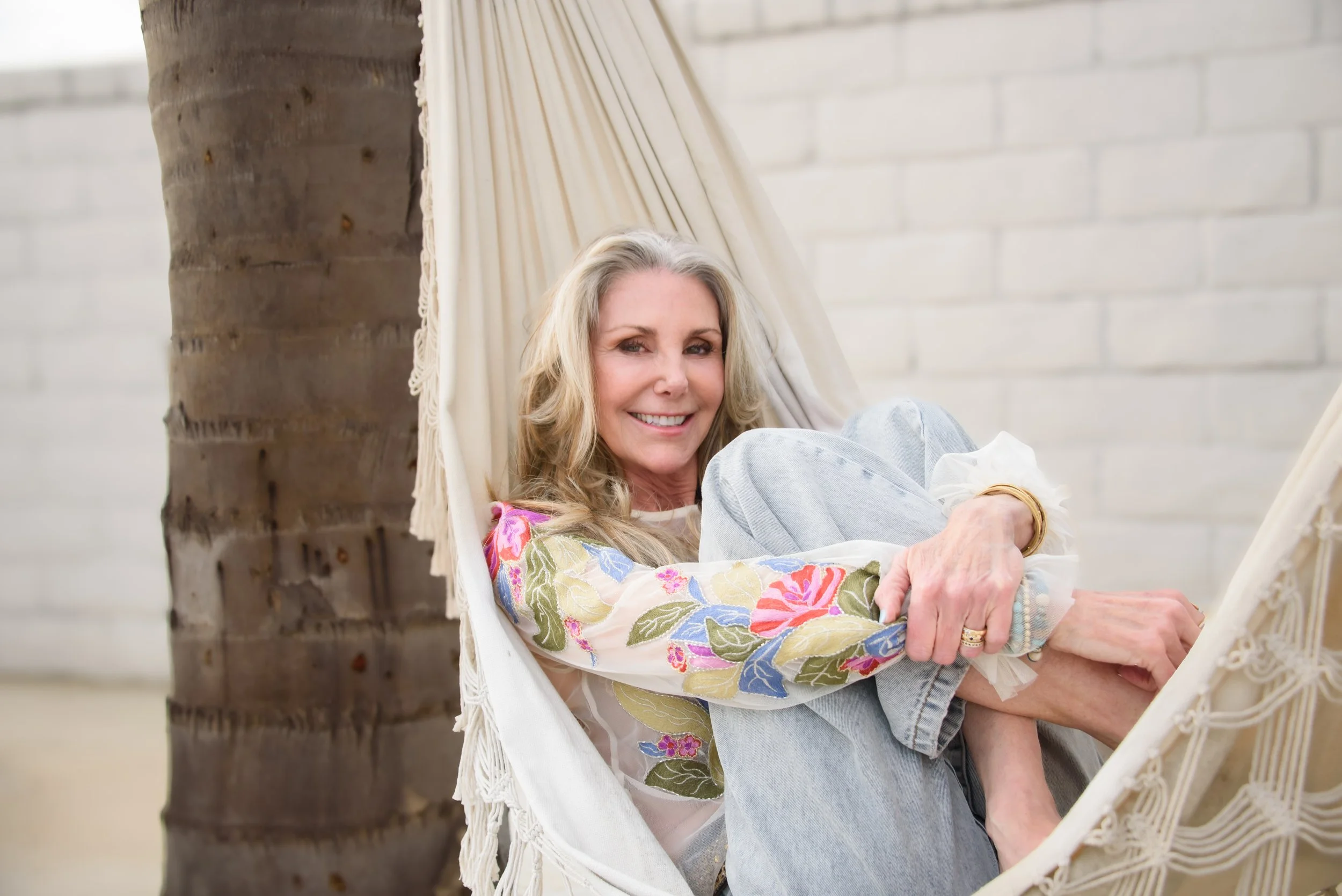 A smiling woman with blonde hair sitting in a hammock near a palm tree, with a white brick wall in the background.