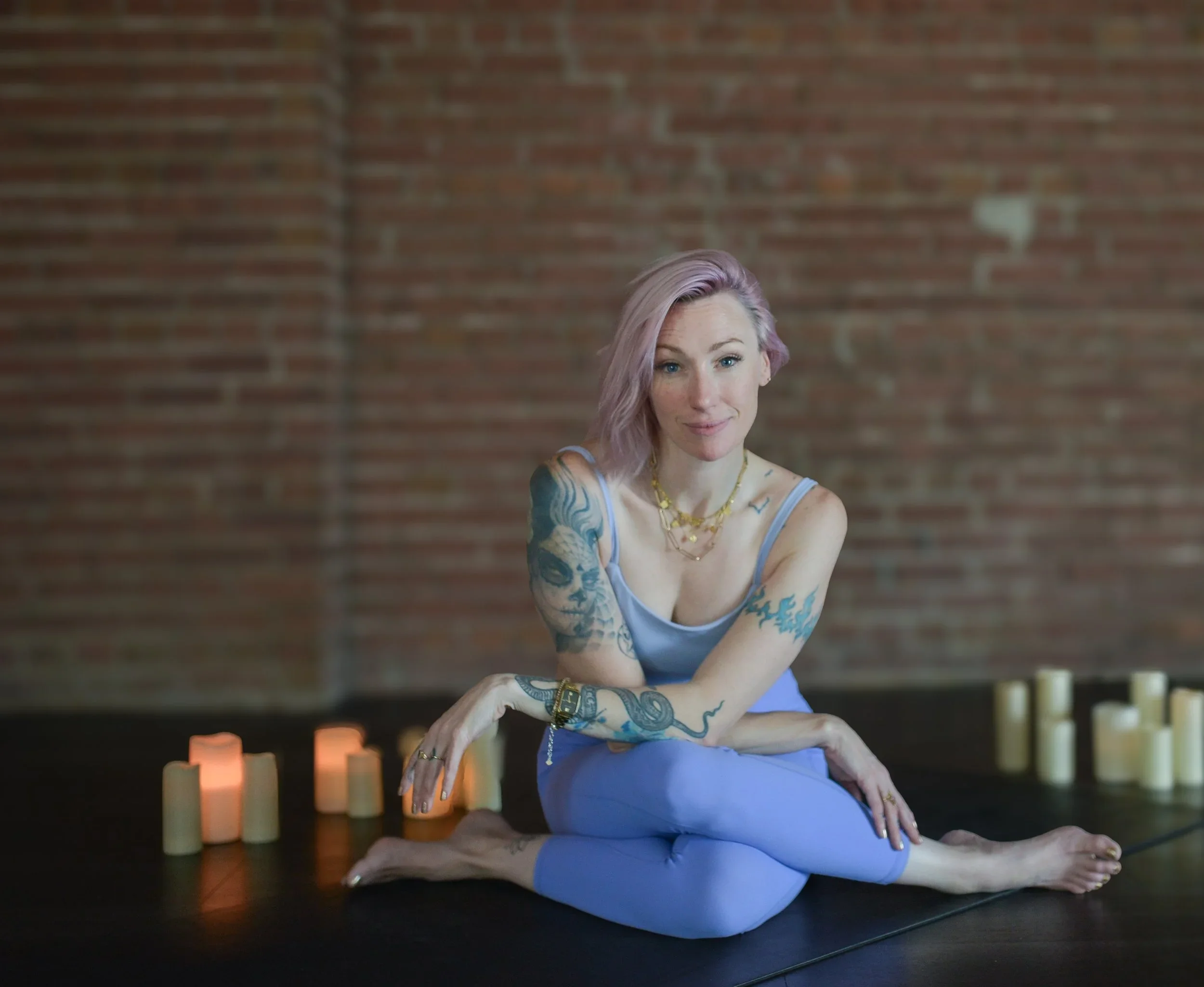 A woman with pastel pink hair practicing yoga on a black mat near candles, in front of a brick wall.