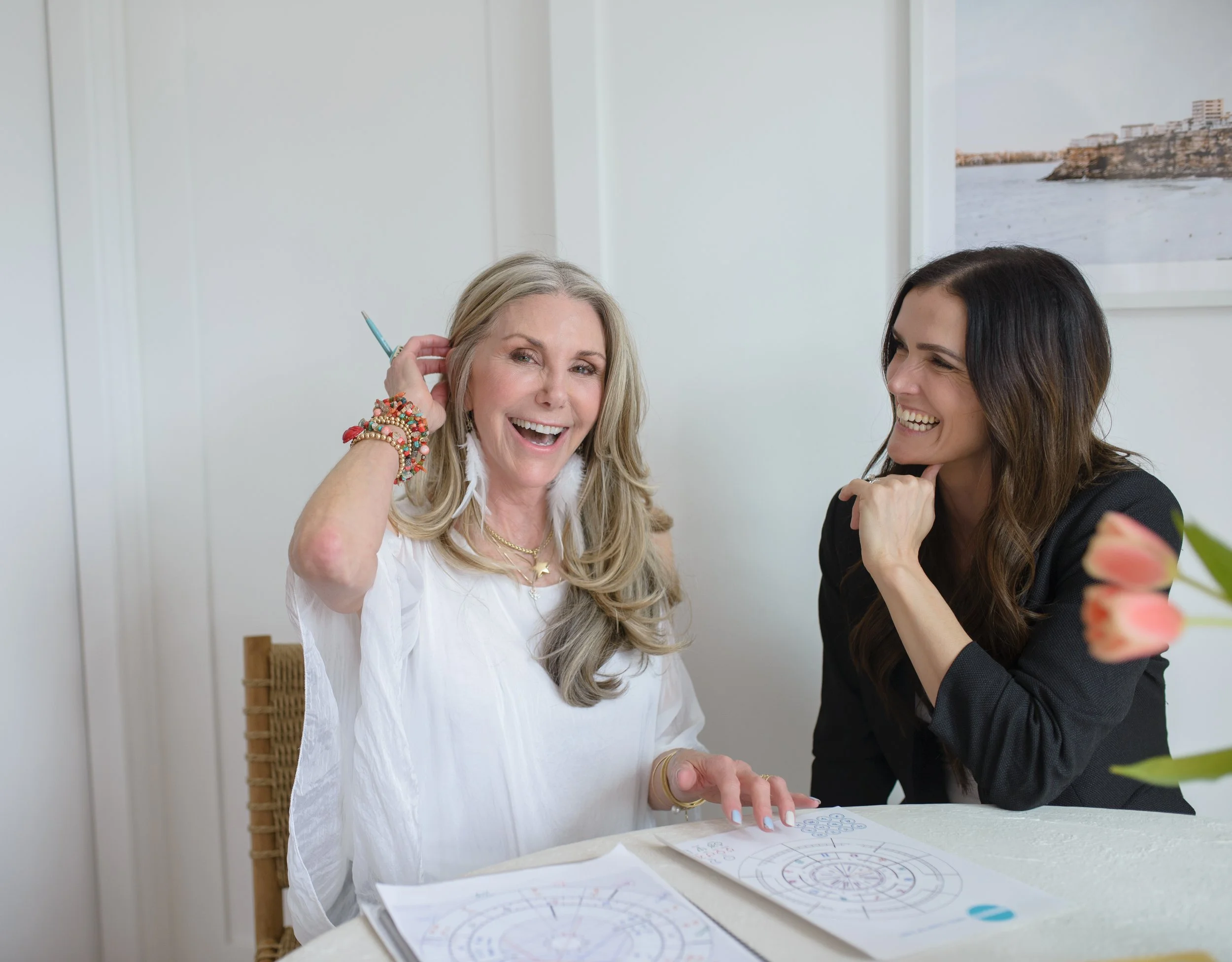 Two women sitting at a table, one with gray hair wearing a white blouse, smiling and holding a pen, the other with dark hair wearing a black blazer, smiling and resting her chin on her hand, having a lively conversation in a bright indoor setting.