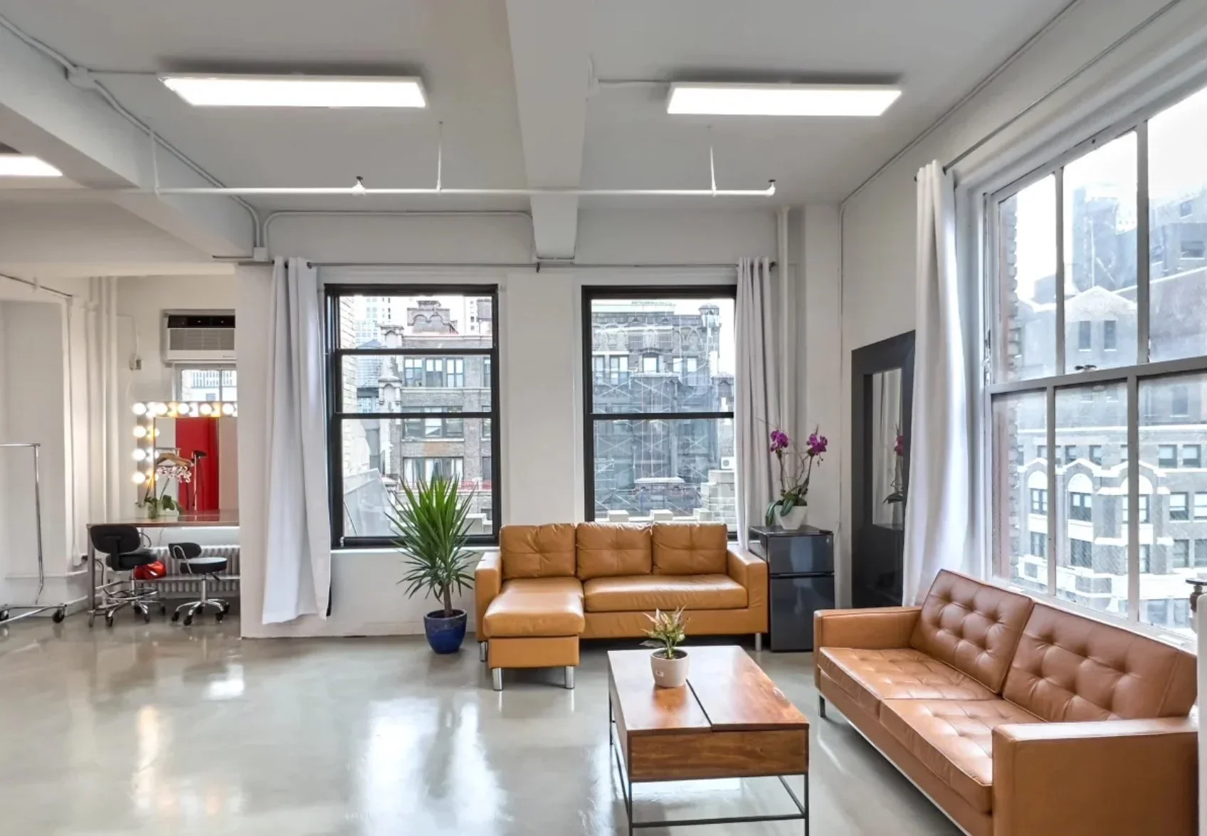 Modern loft apartment with large windows, tan leather sofas, potted plants, a coffee table, a makeup mirror, and bright lighting.