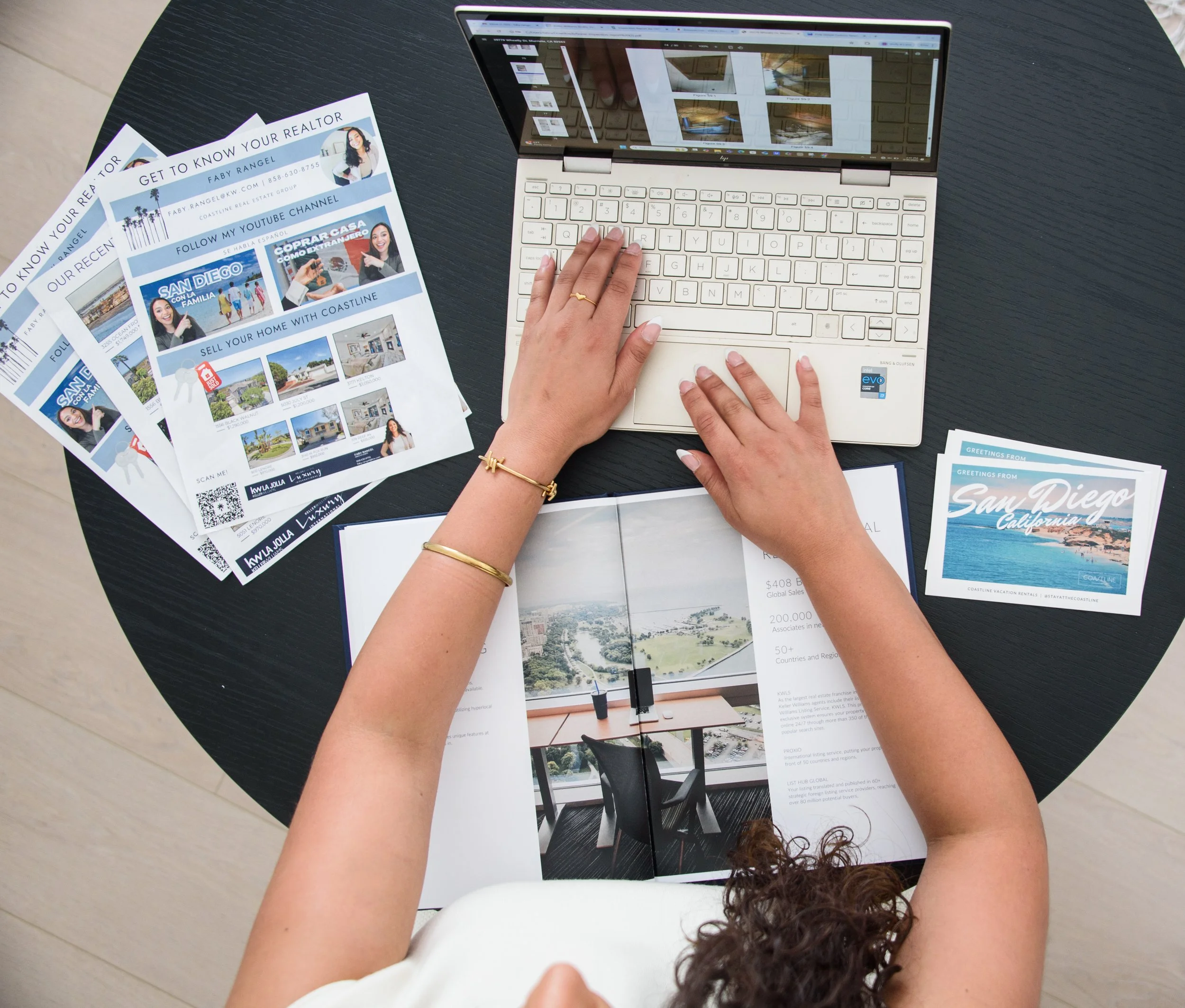 A person working on a laptop at a round black table, with brochures and flyers about San Diego, California, and real estate, along with an open magazine showing interior design.