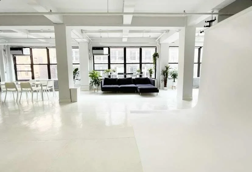 Bright, modern office space with large windows, a black sofa, potted plants, and a white dining area with chairs.
