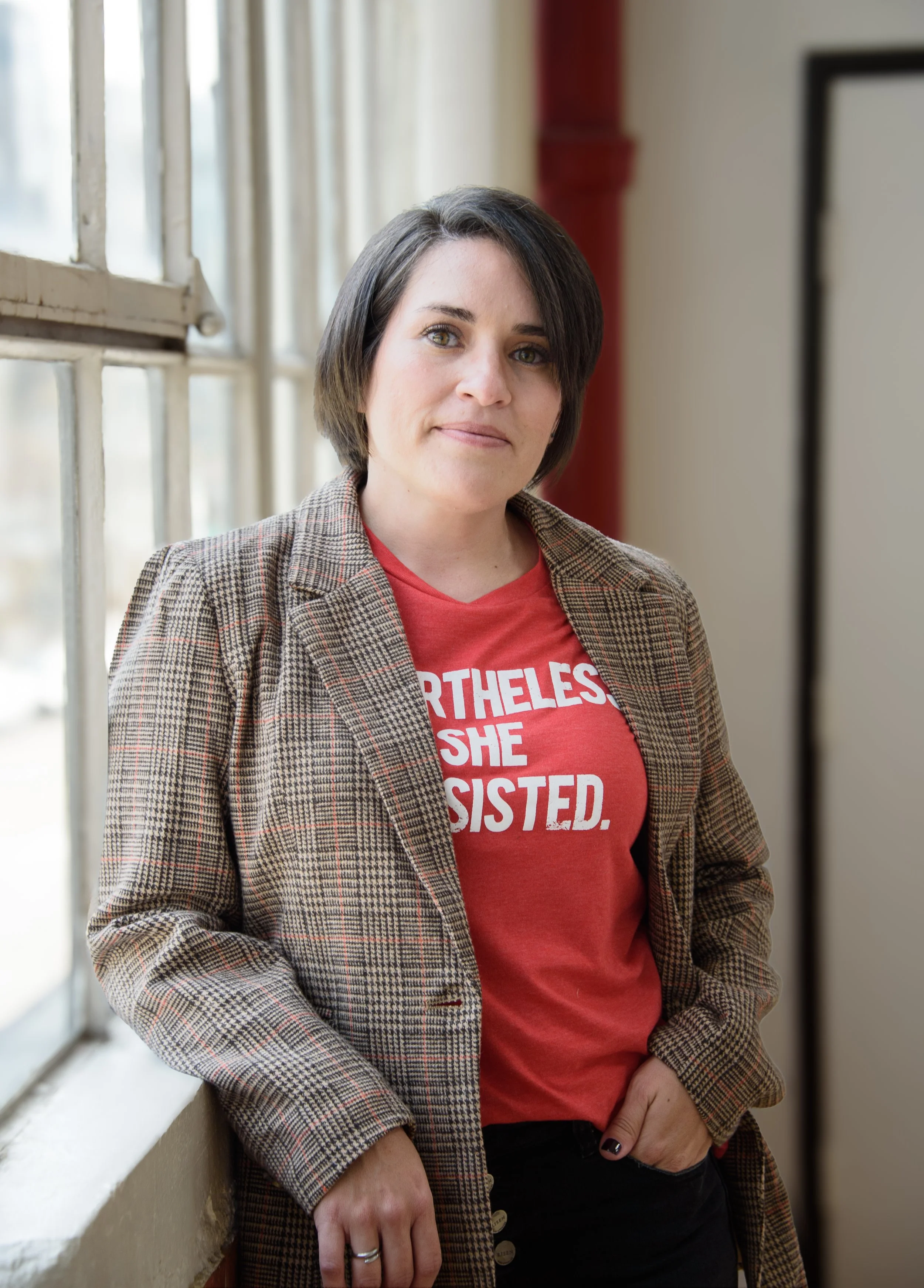 A woman with short dark hair wearing a red t-shirt with white text and a plaid blazer, standing indoors near a window.