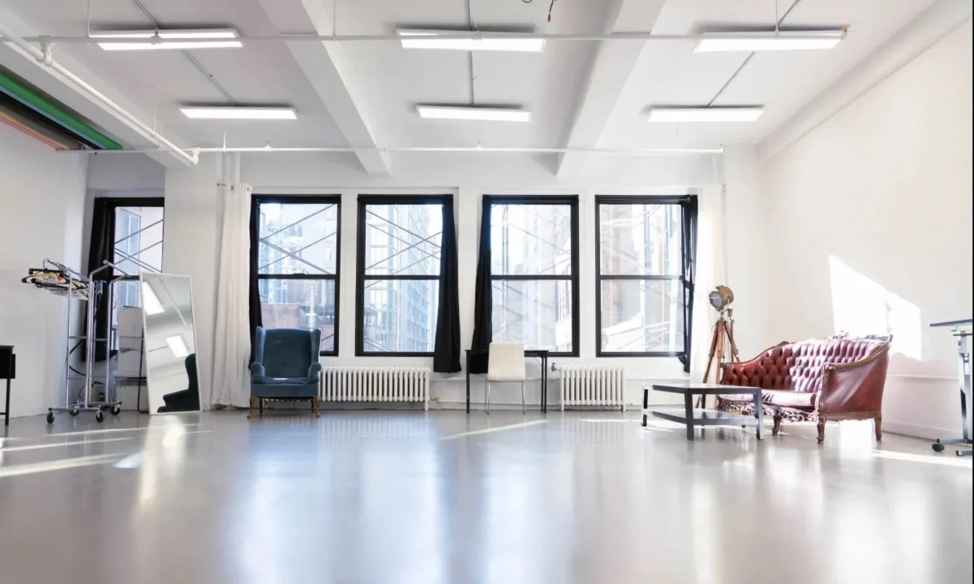 An empty spacious room with large windows, a vintage red sofa, a blue armchair, a white chair, a mirror, and various studio equipment.