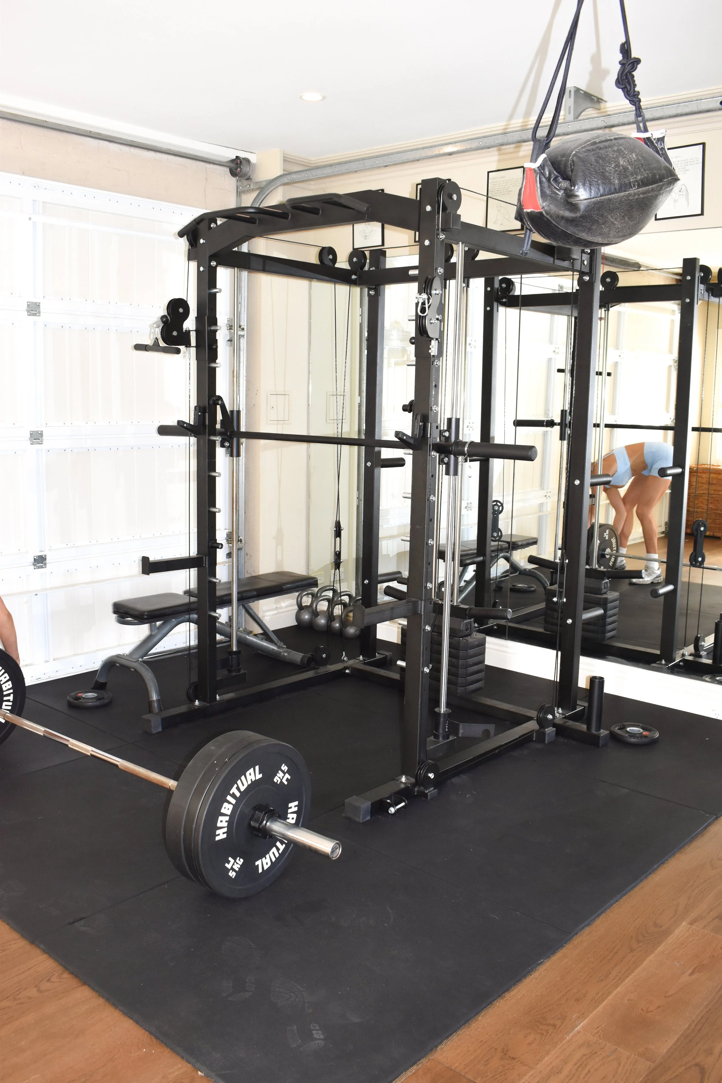 Weightlifting equipment in a gym, including a barbell with weights on the floor, a power rack, a punching bag hanging from the ceiling, and a woman in workout clothes bending over in the background.