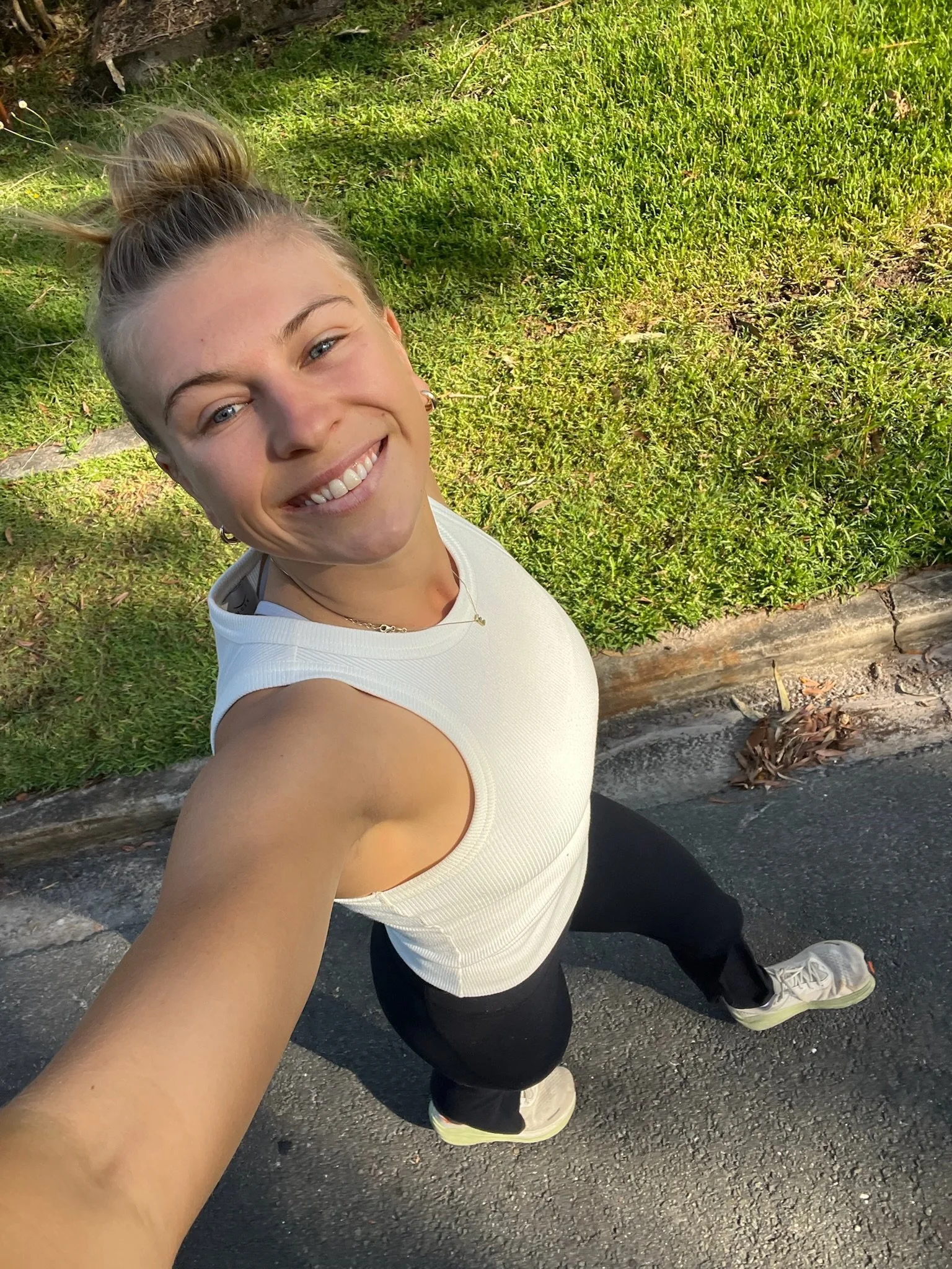 A woman smiling, taking a selfie outdoors on a sunny day, wearing a white sleeveless top, black pants, and sneakers, standing on a sidewalk next to a grassy area.