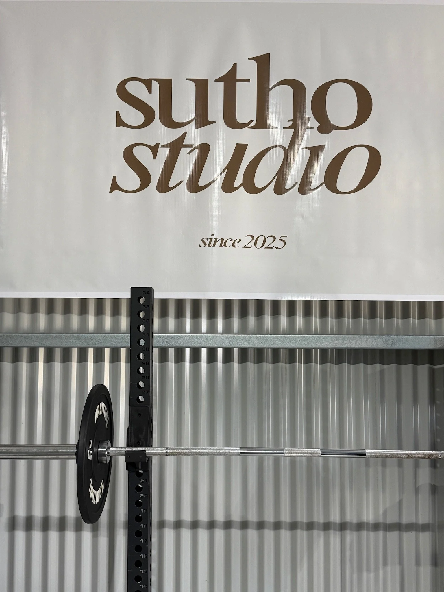 A barbell with a weight plate mounted on it in front of a metallic garage door. A sign above reads 'sutho studio since 2025'.