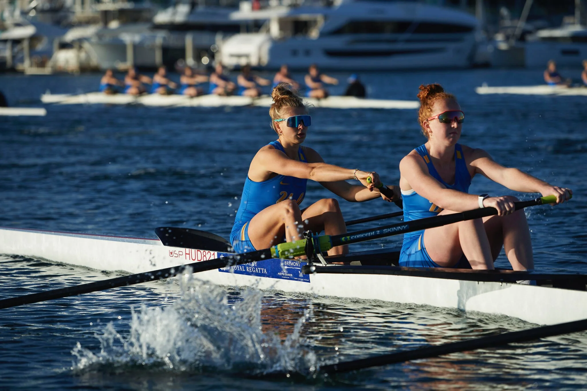 Two women in blue athletic outfits rowing a boat on the water, with a lineup of passing boats and yachts in the background.