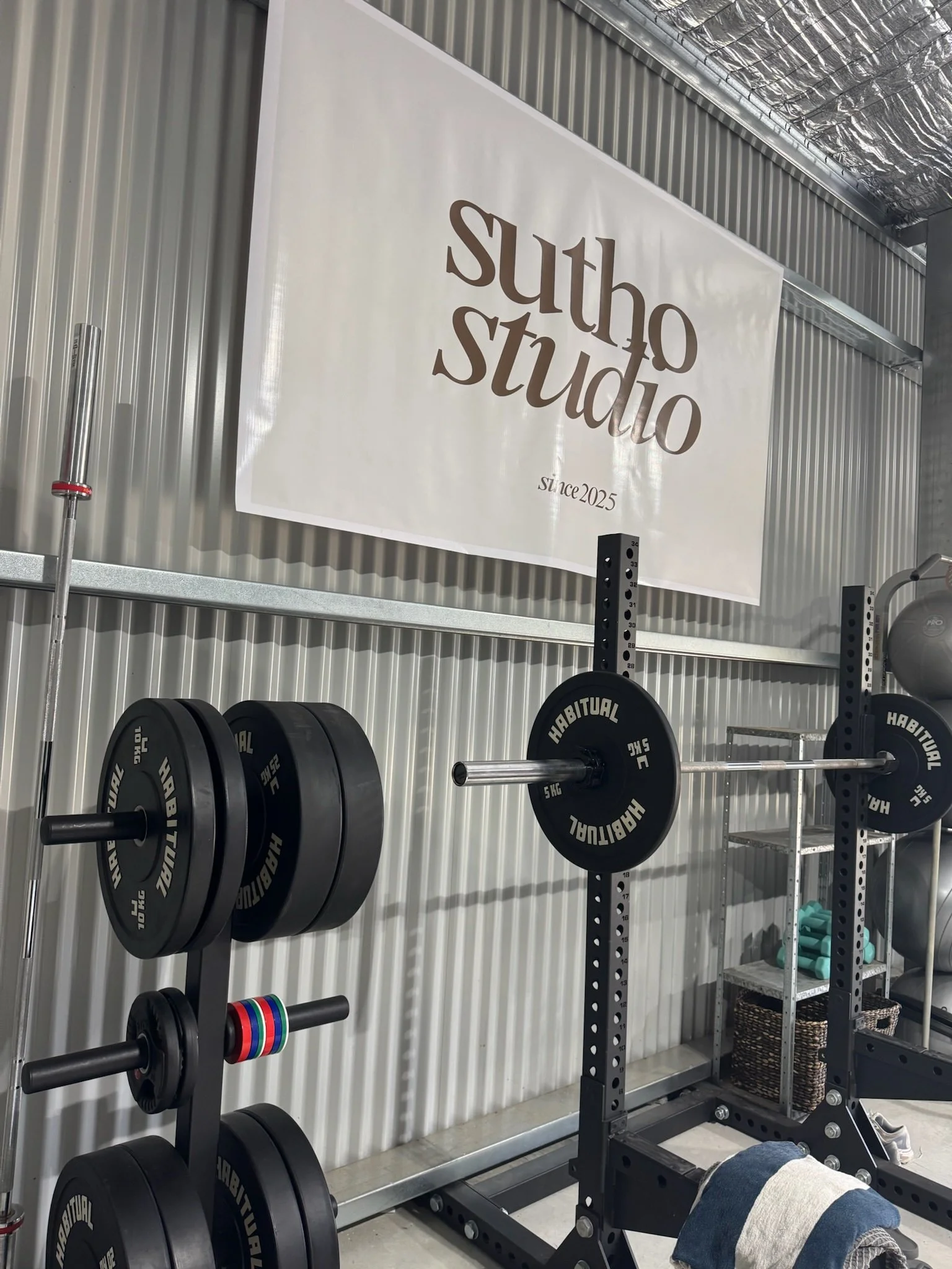 Interior of a gym with weightlifting equipment, a barbell with weights, a rack of weights, a banner reading 'sutho studio since 2025', and shelving with workout gear.