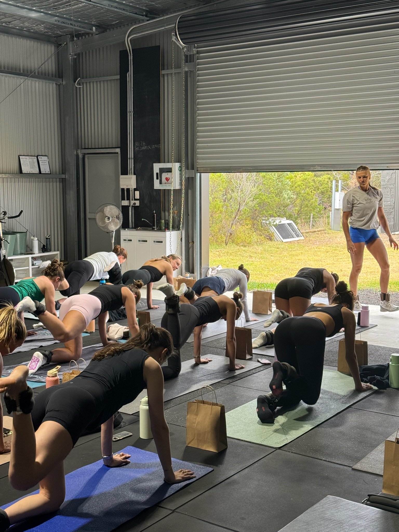 Group of women participating in a fitness class inside a gym with an open garage door showing outdoor greenery.