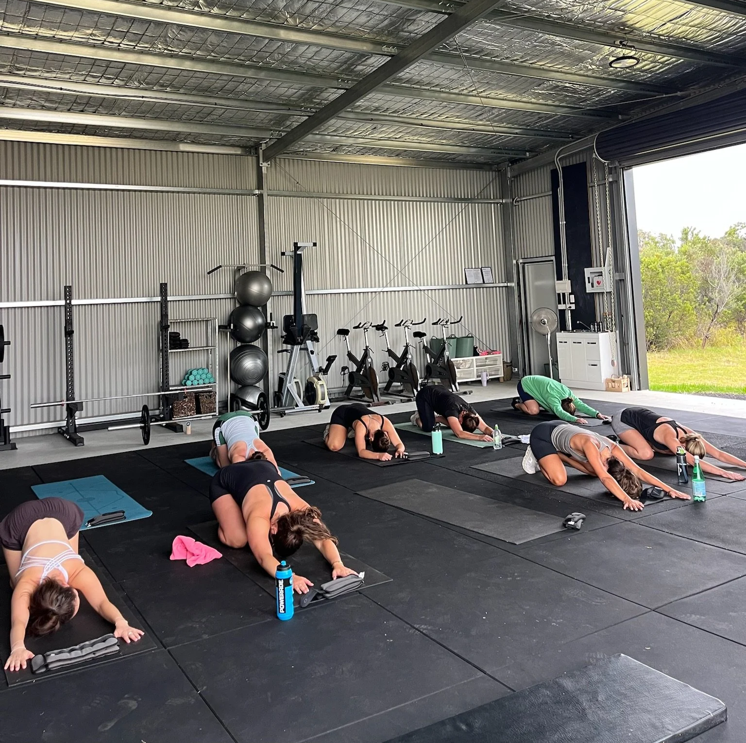 People participating in a yoga class inside a spacious garage or industrial-style building, practicing the child's pose on black mats.