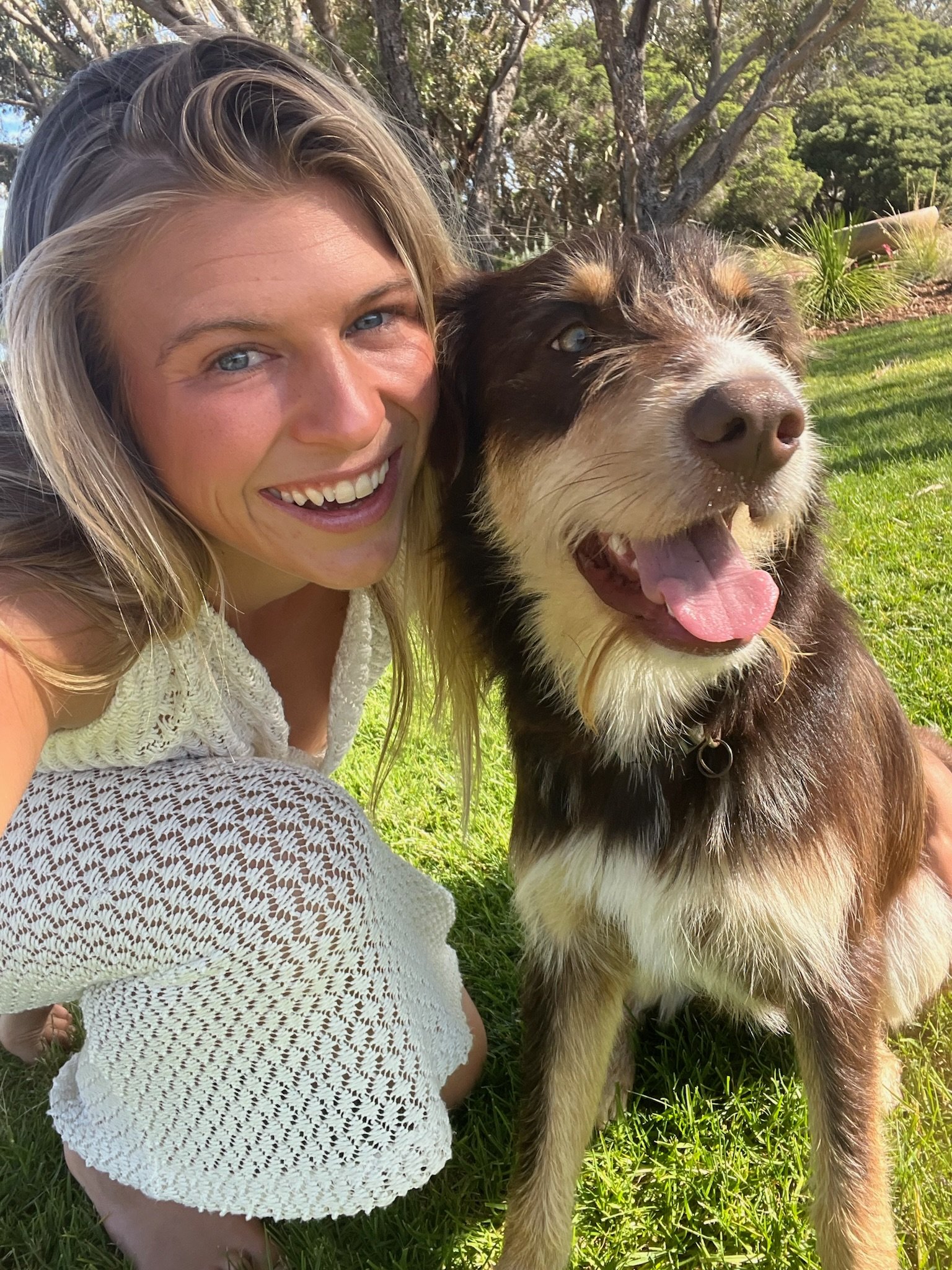 A woman with blonde hair and a white lace top taking a selfie outdoors with a happy brown and white Australian Shepherd dog on a sunny day in a grassy park.