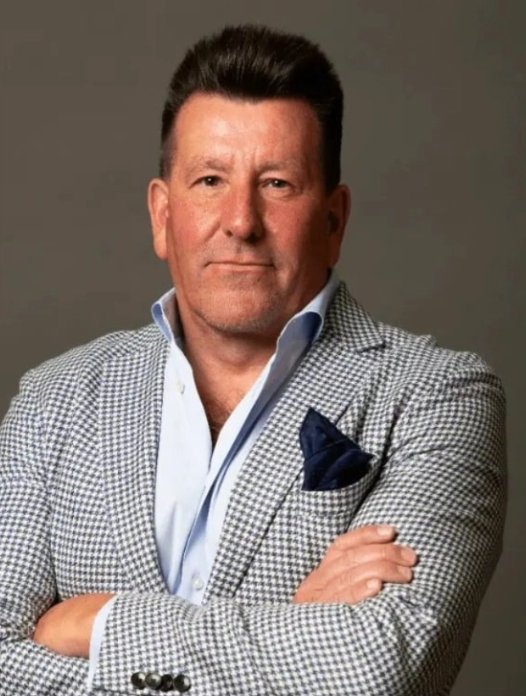 A man with dark hair, wearing a checkered blazer, white shirt, and navy pocket square, standing with arms crossed against a gray background.