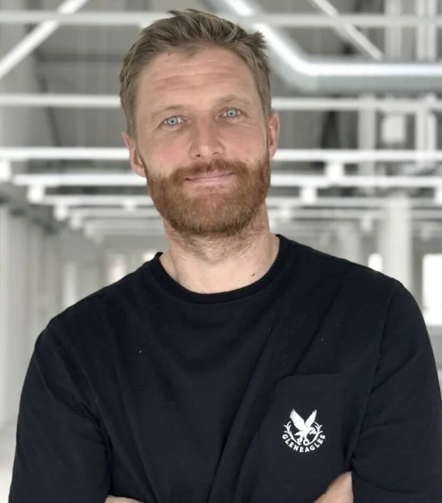 A man with curly dark hair, glasses, and a beard, looking to the side, sitting indoors with a neutral background and a plant in the background.