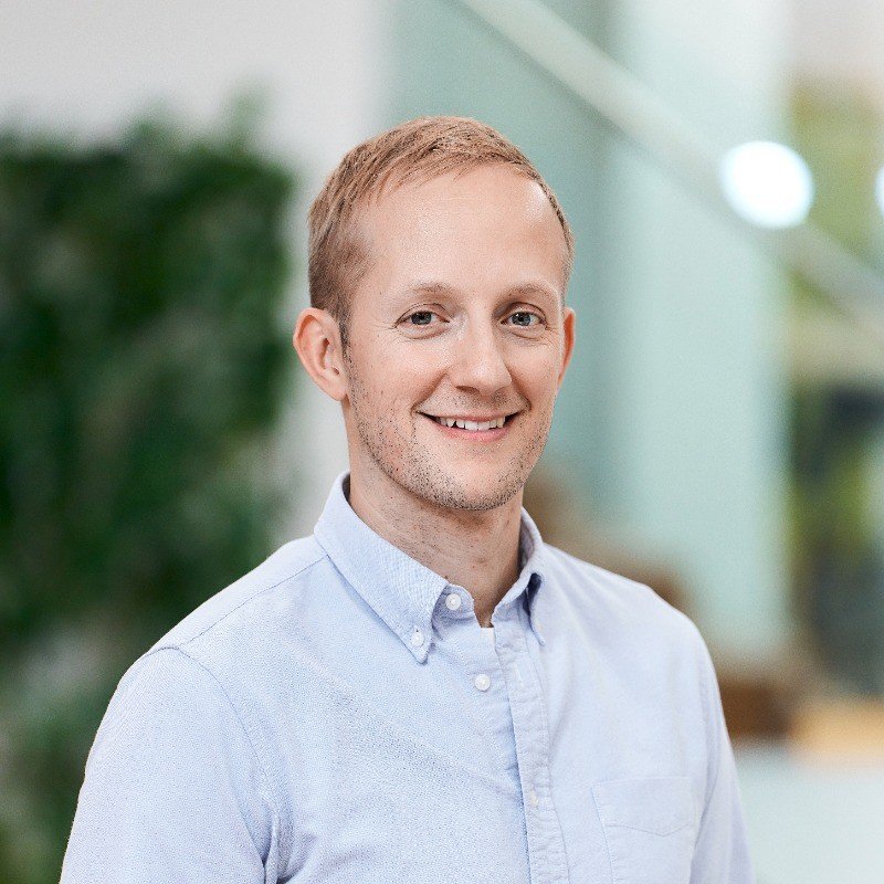 A young man with short red hair and fair skin, smiling, wearing a light blue button-up shirt, standing indoors with blurred background of greenery and light.