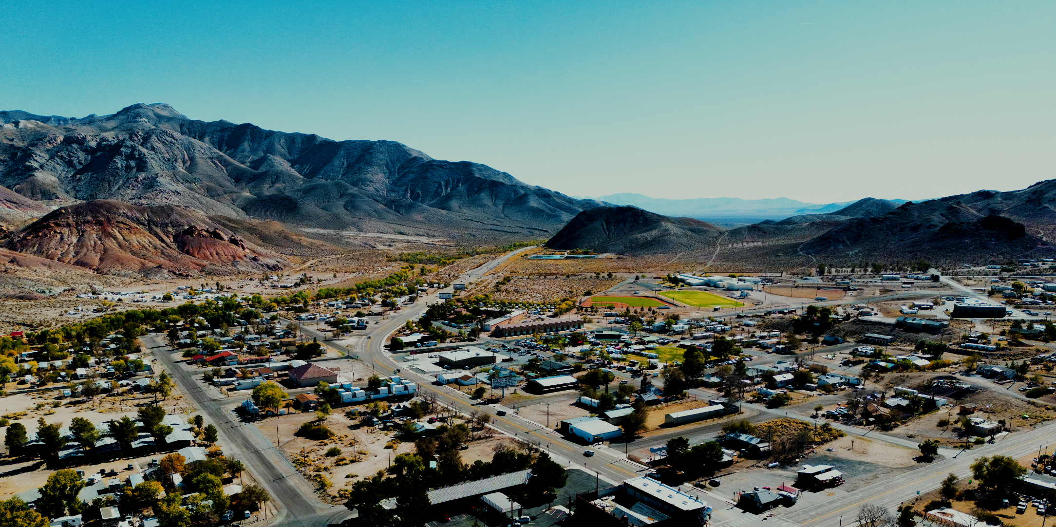 Aerial view of Beatty, Nevada with mountains in the background, showing streets, buildings, trees, and a sports field.