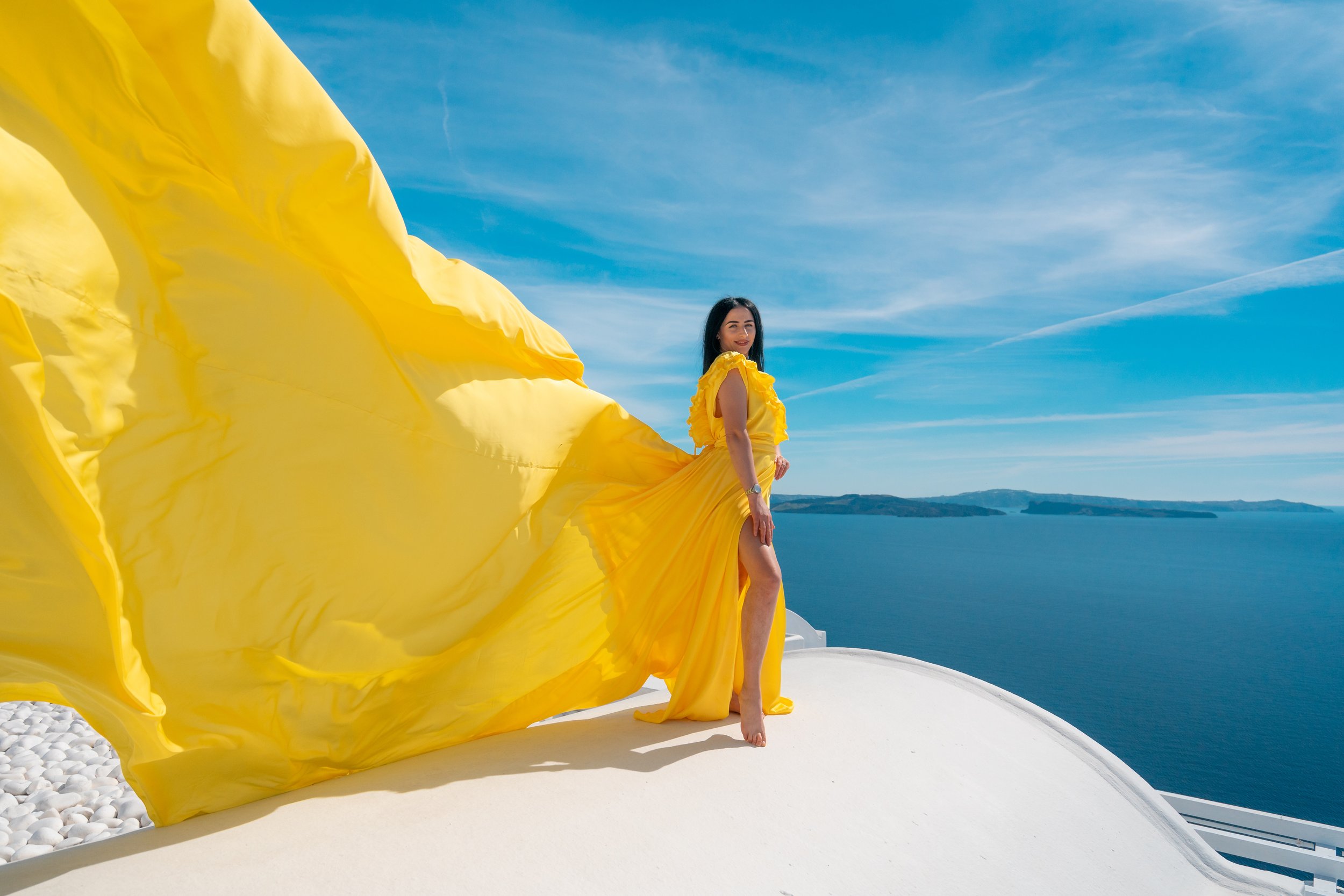 A woman in a long yellow dress standing on a white surface outside, with the sea and hills in the background, under a blue sky. The dress has a flowing train that billows behind her.