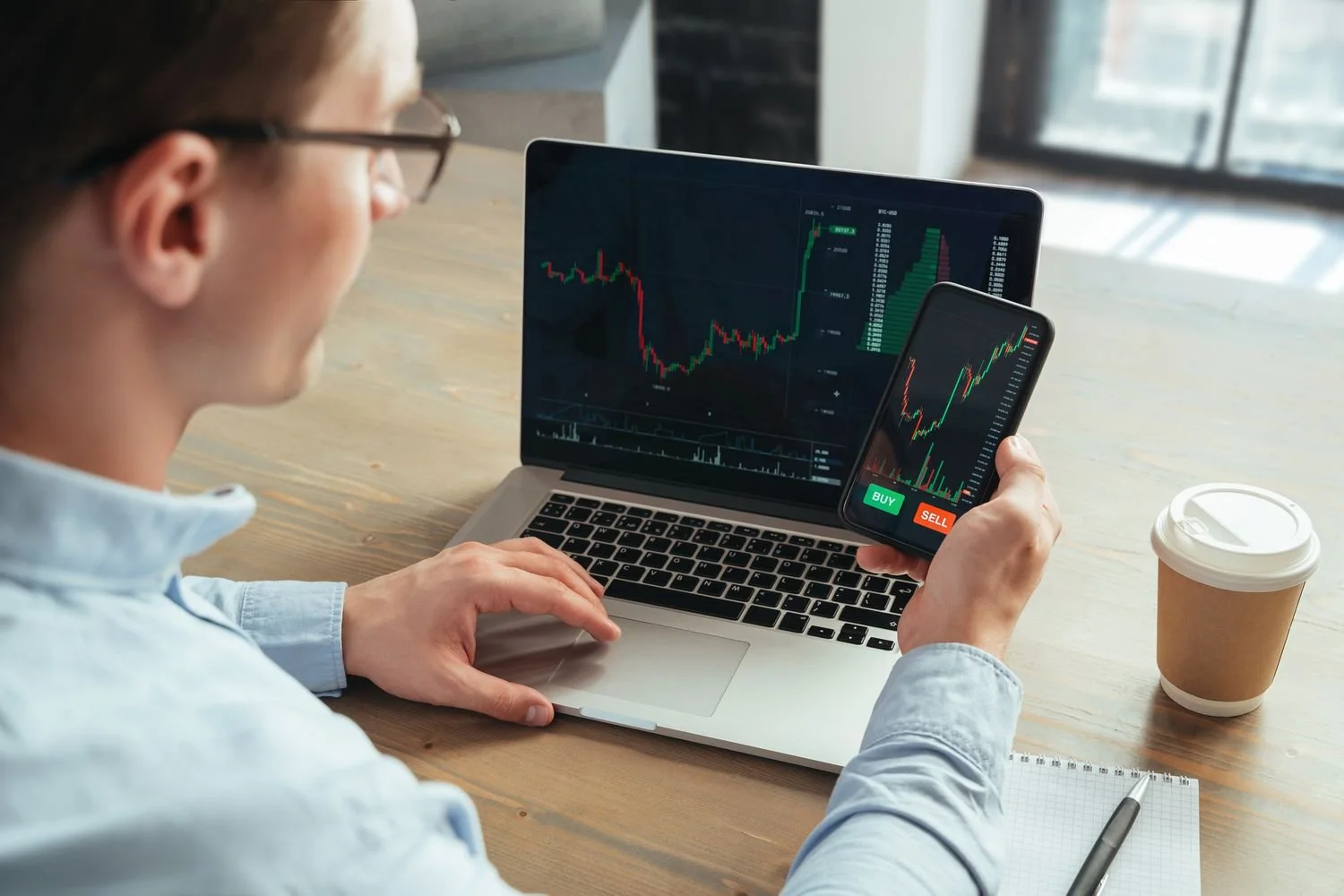 Person looking at stock trading charts on a laptop and smartphone, with a notebook and coffee on a wooden table.