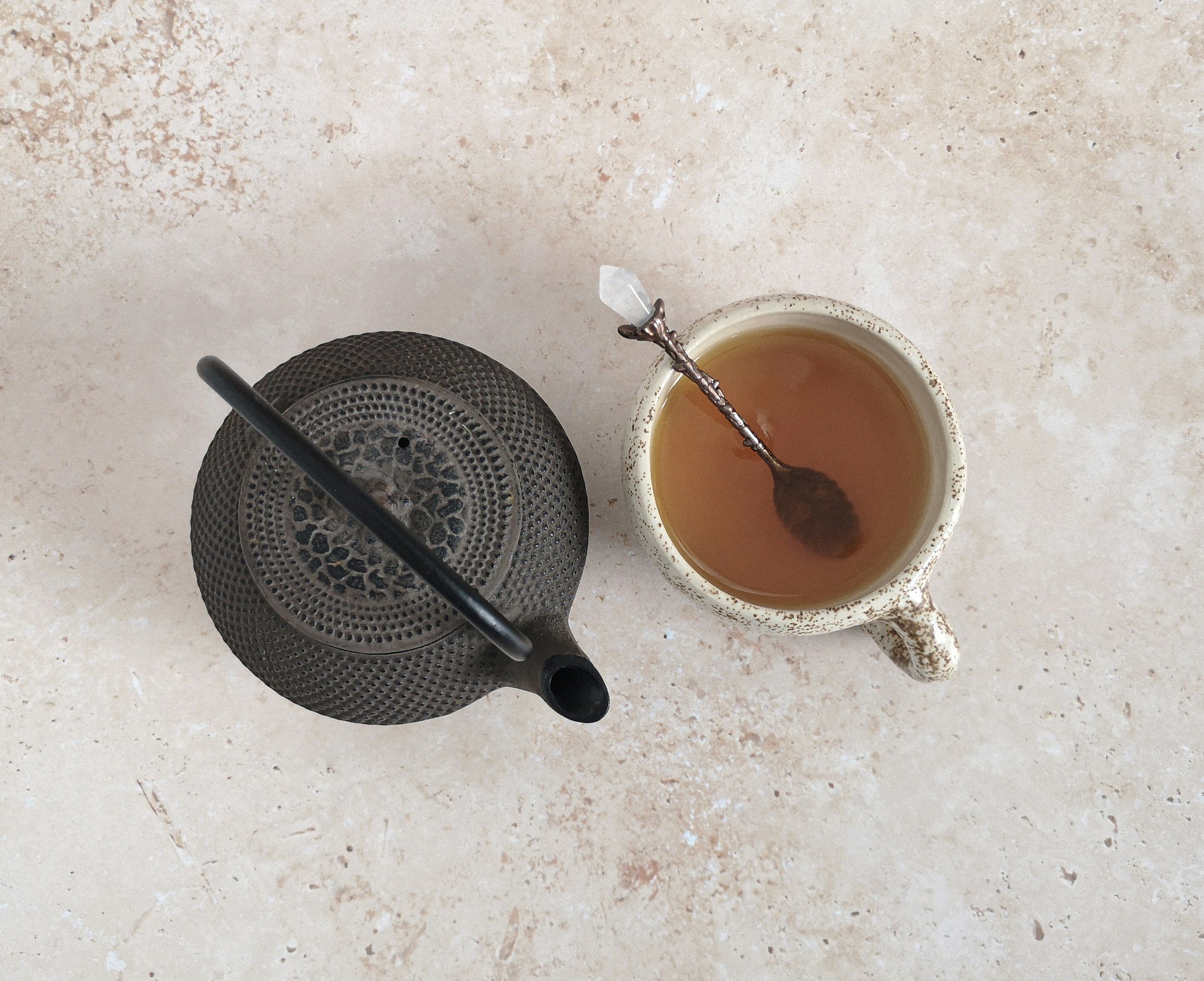 Overhead view of a textured teapot, a mug filled with tea with a silver spoon, and a black straw on a beige surface.