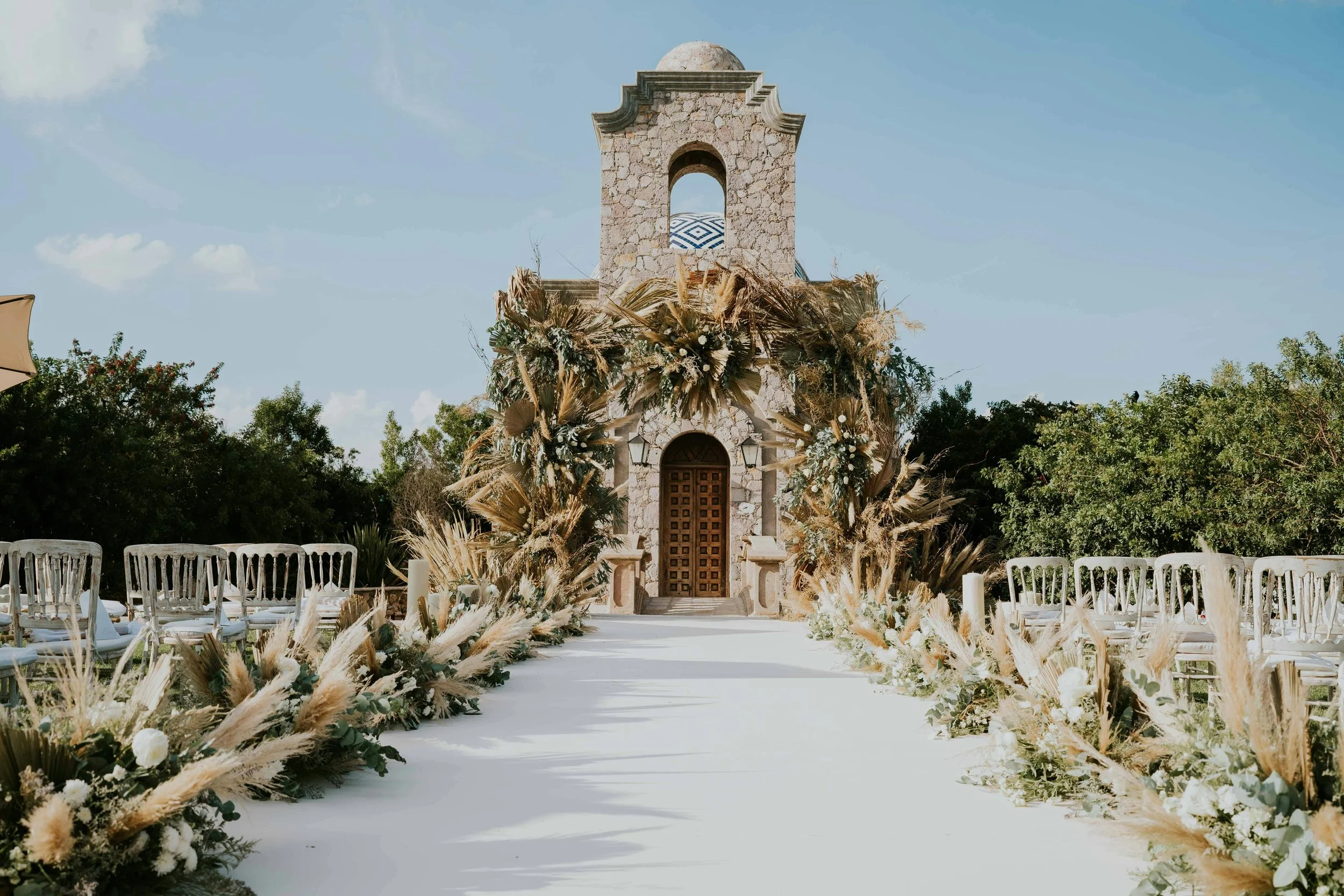 Outdoor wedding setup with white chairs and floral arrangements on a white aisle, leading to a stone chapel with floral decorations and a blue sky background.