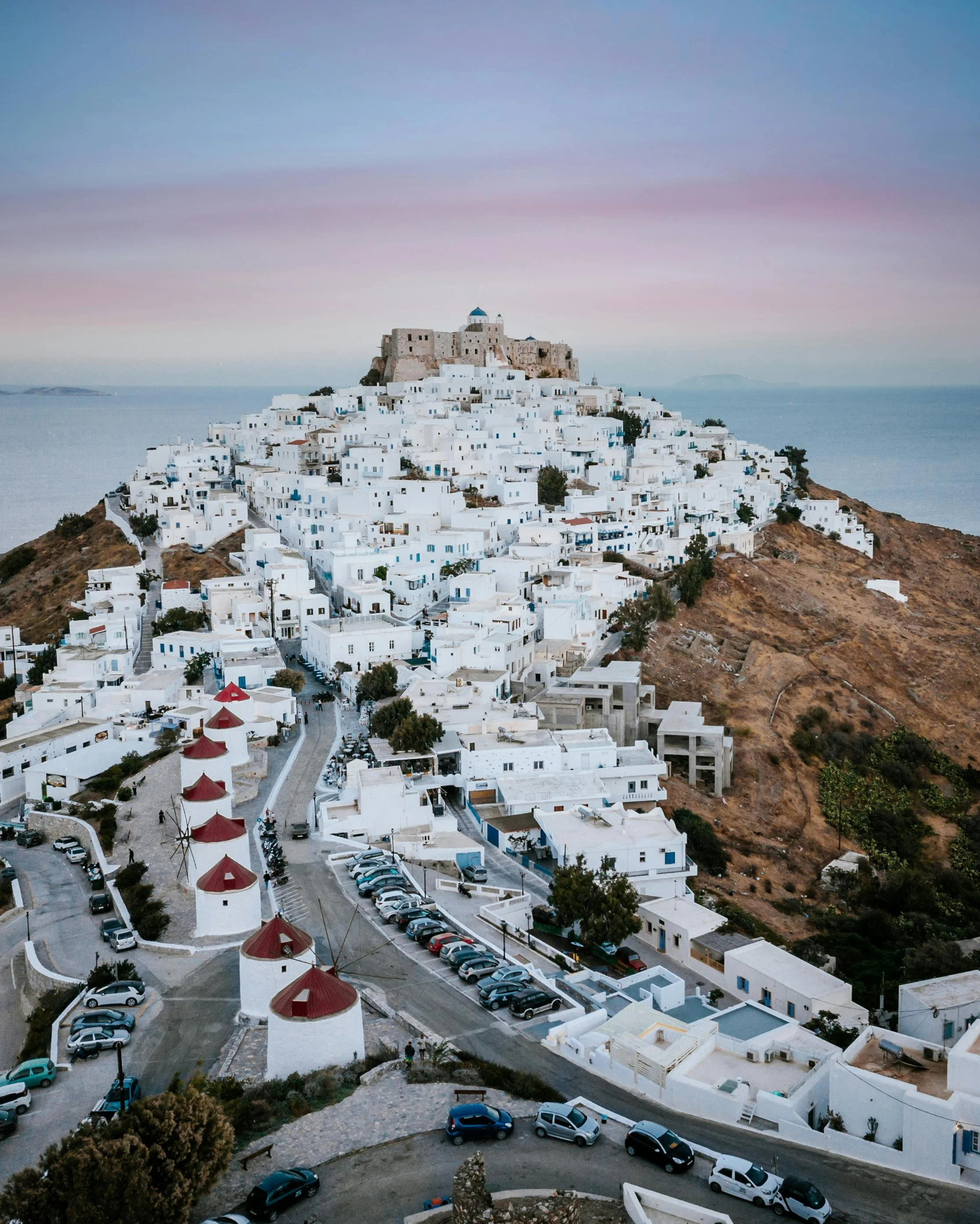 Aerial view of white buildings with red windmills in the foreground and a hillside town on a hilltop in Greece.