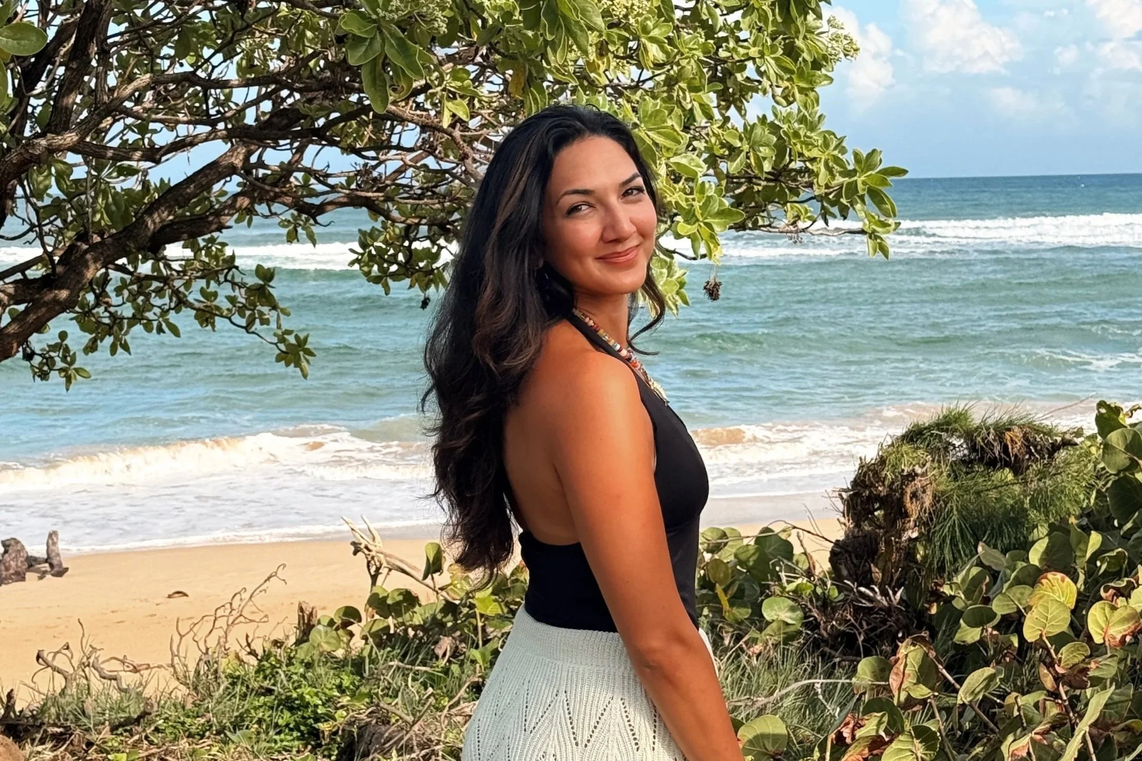 A woman with long dark hair smiling at the camera while standing on a beach with ocean waves in the background. She is near greenery and trees.