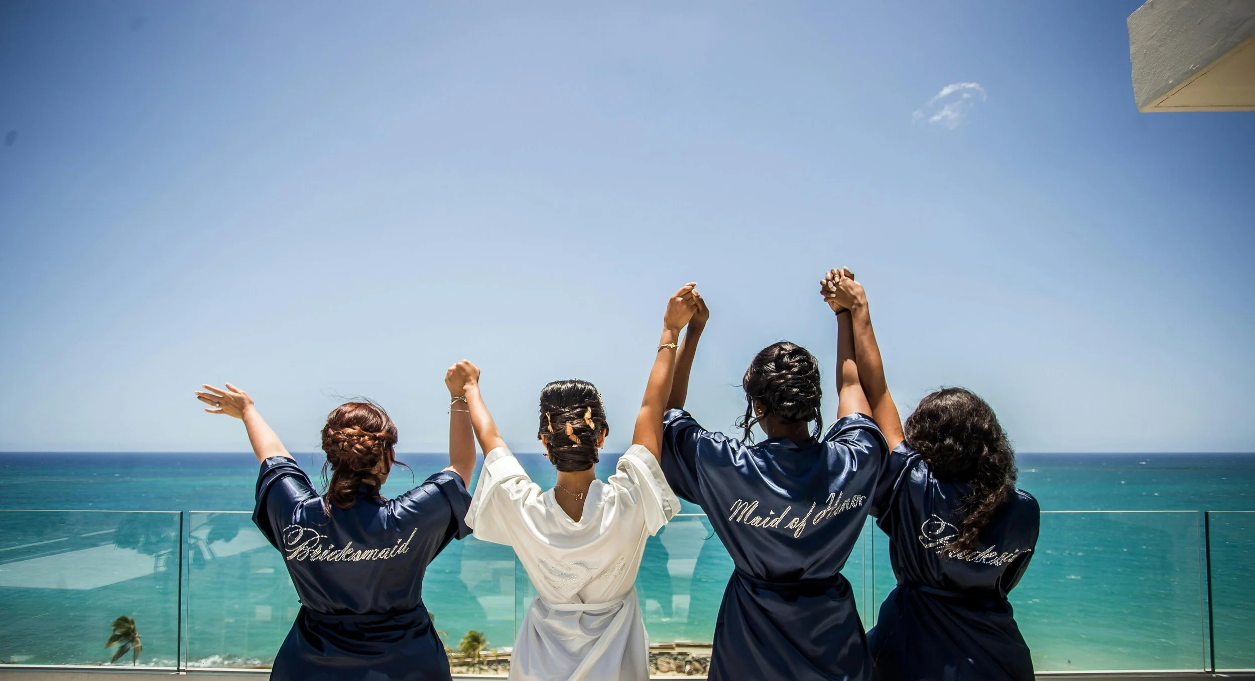 Four women in satin robes with titles like 'Bridemaid,' 'Maid of Honor,' and 'Bridesmaid' stand on a balcony overlooking the ocean, holding hands up in celebration.