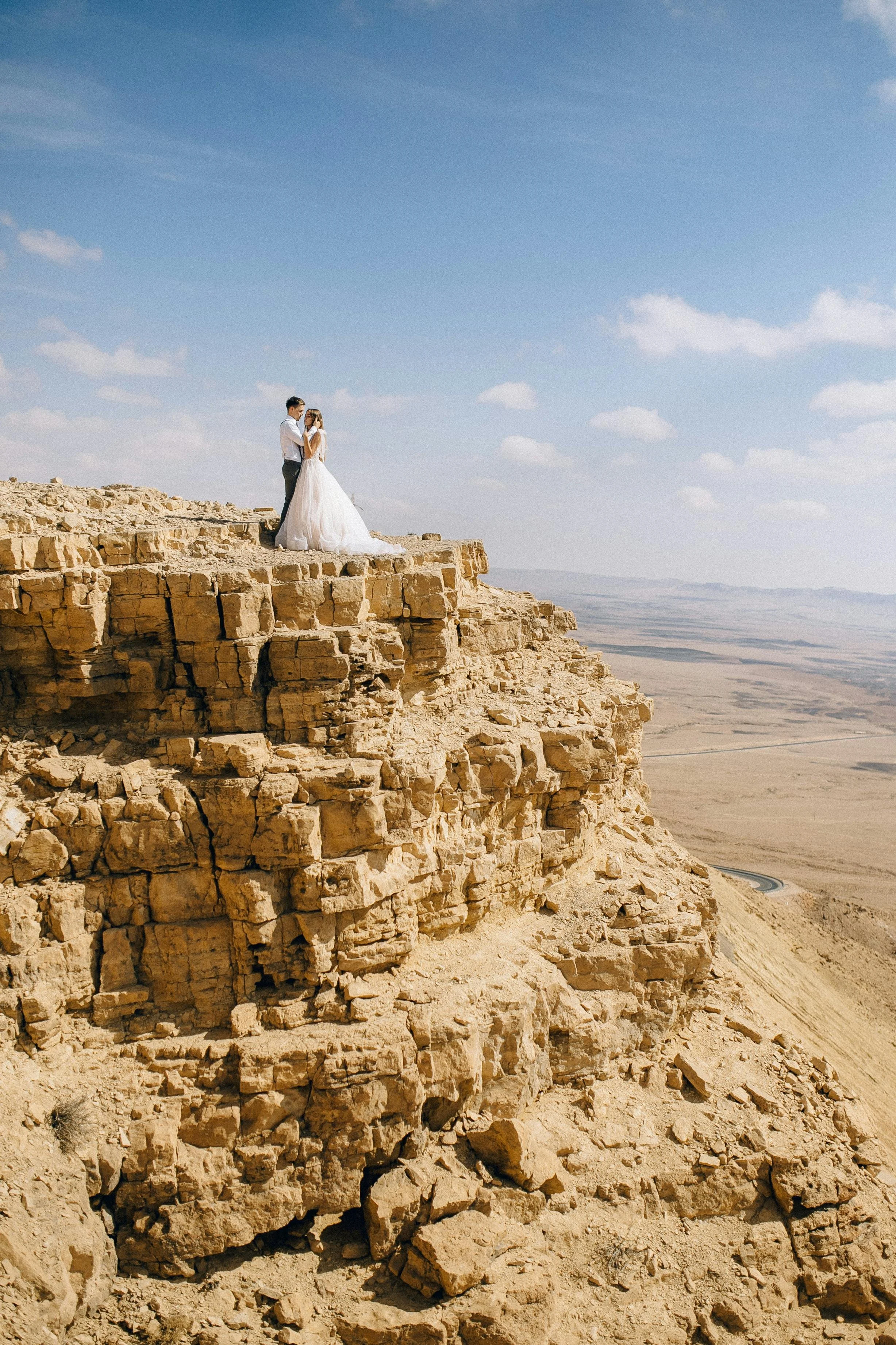 A couple in wedding attire stands on a rocky edge of a desert cliff under a blue sky with scattered clouds, holding hands and facing each other.